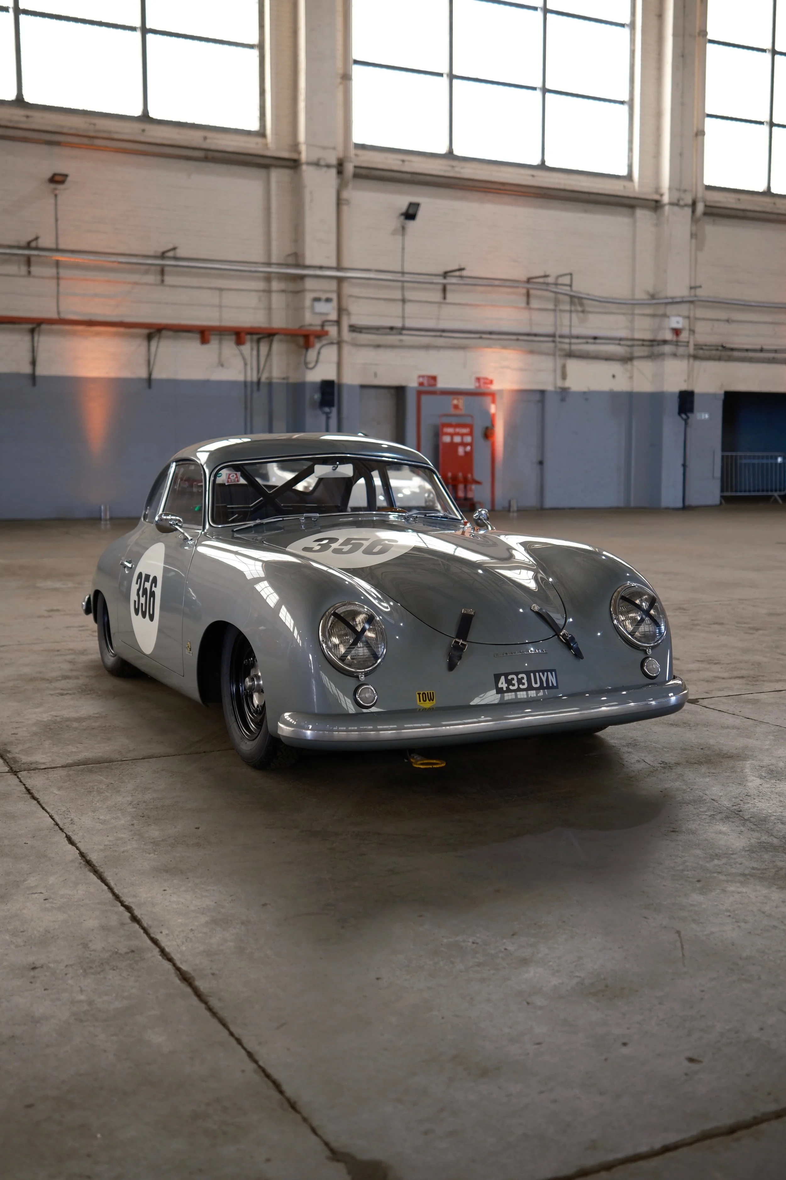 A vintage silver racing car with the number 356 painted on its hood and doors, parked inside an industrial-style building with large windows and exposed piping.