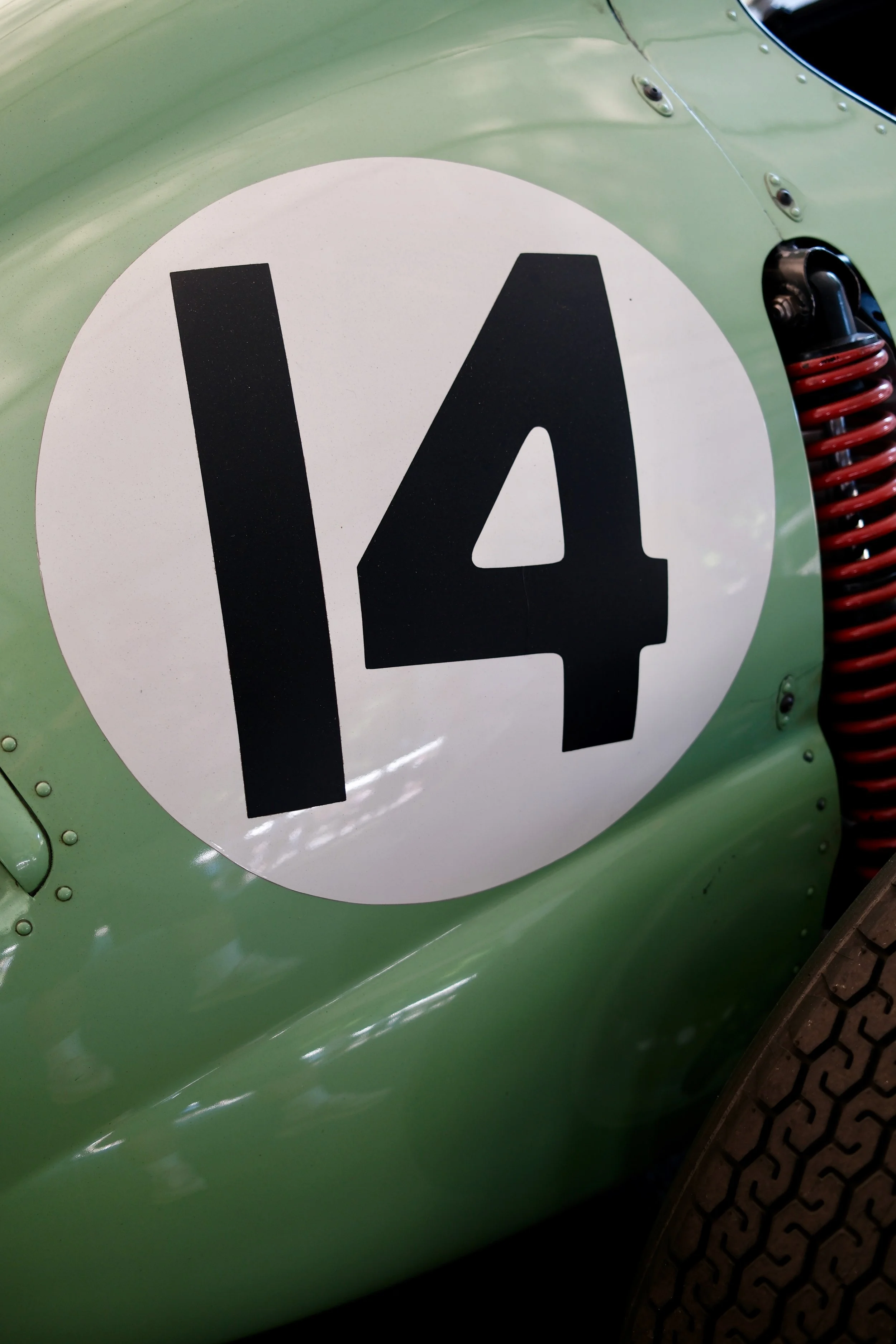 Close-up of a green vehicle body with a white circular sticker displaying the number 14 in black, a red coil spring suspension component, and part of a black tire with a textured tread pattern.