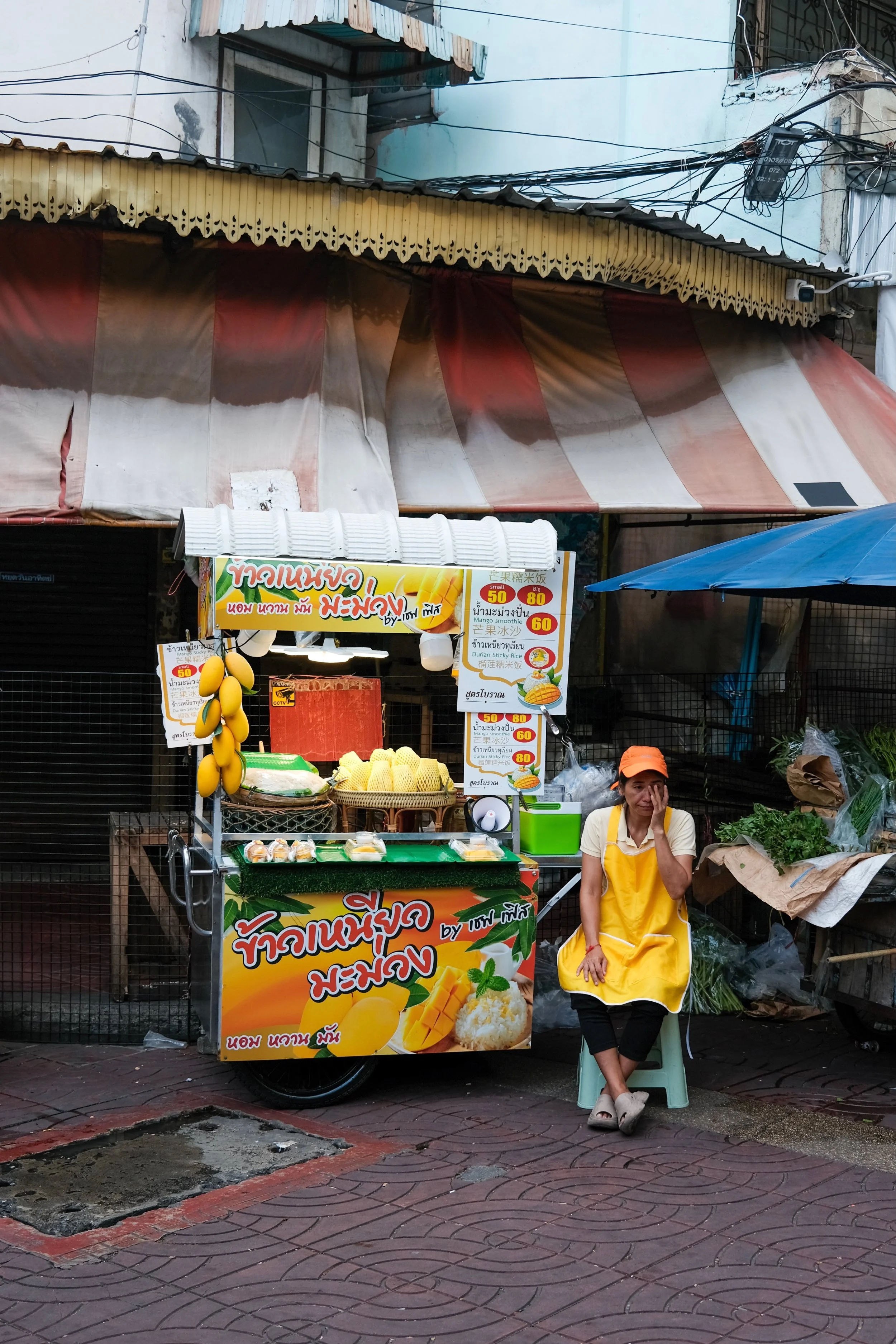 A street vendor stand selling mango sticky rice, with a woman sitting on a small stool in yellow apron, holding her face. The stand has mangoes, rice, and a colorful sign with Thai and Chinese writing promoting the dish, located on a sidewalk in an u