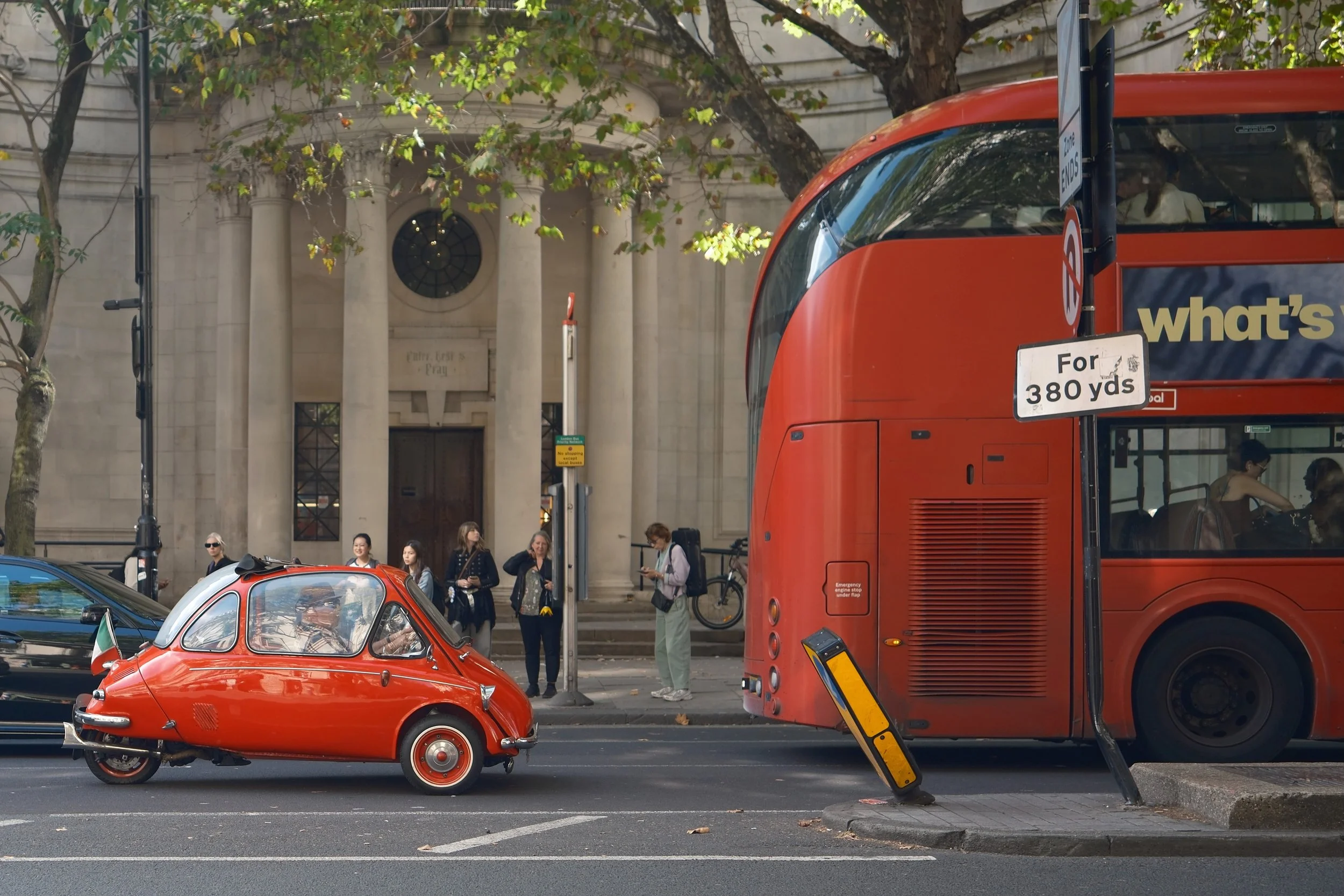 Red double-decker bus and a small red car on a city street with pedestrians and trees in the background.