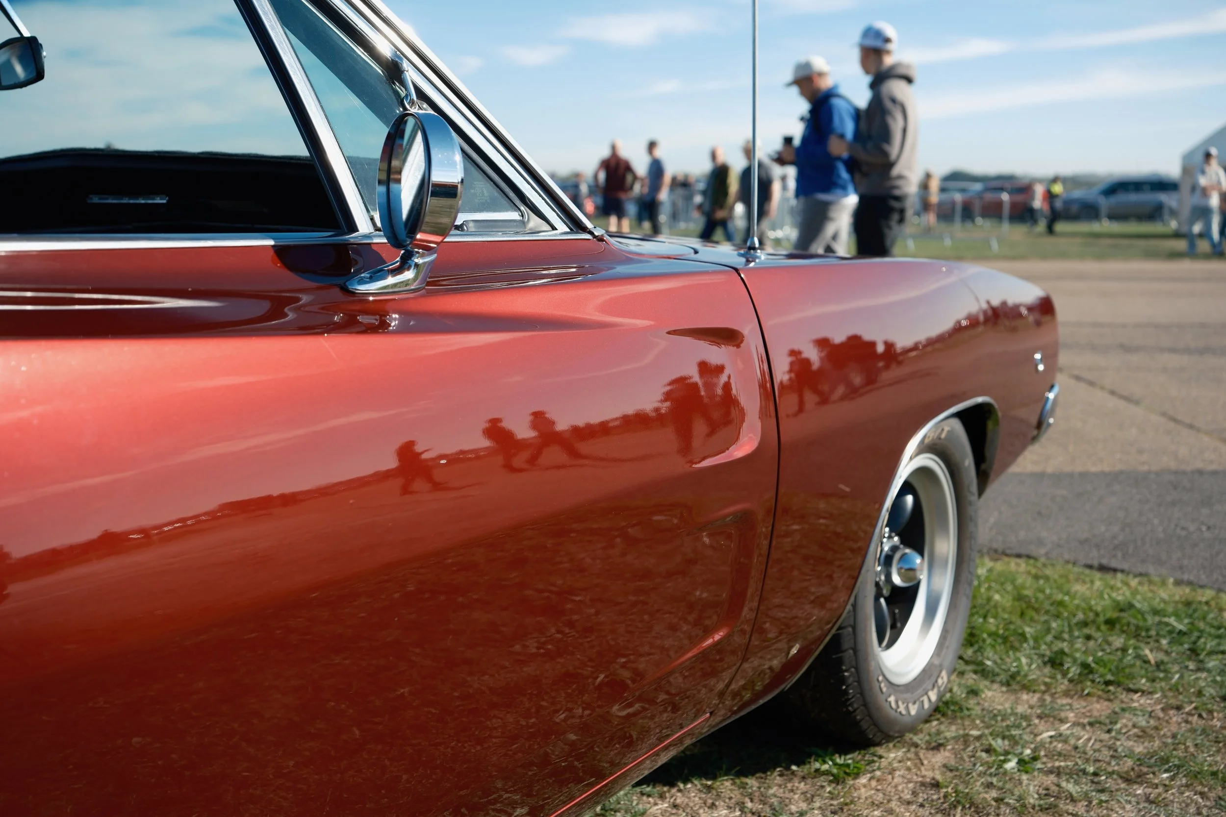 Close-up of a vintage red car at an outdoor event, with a focus on the side mirror and body, reflecting other cars and people in the background on a sunny day.