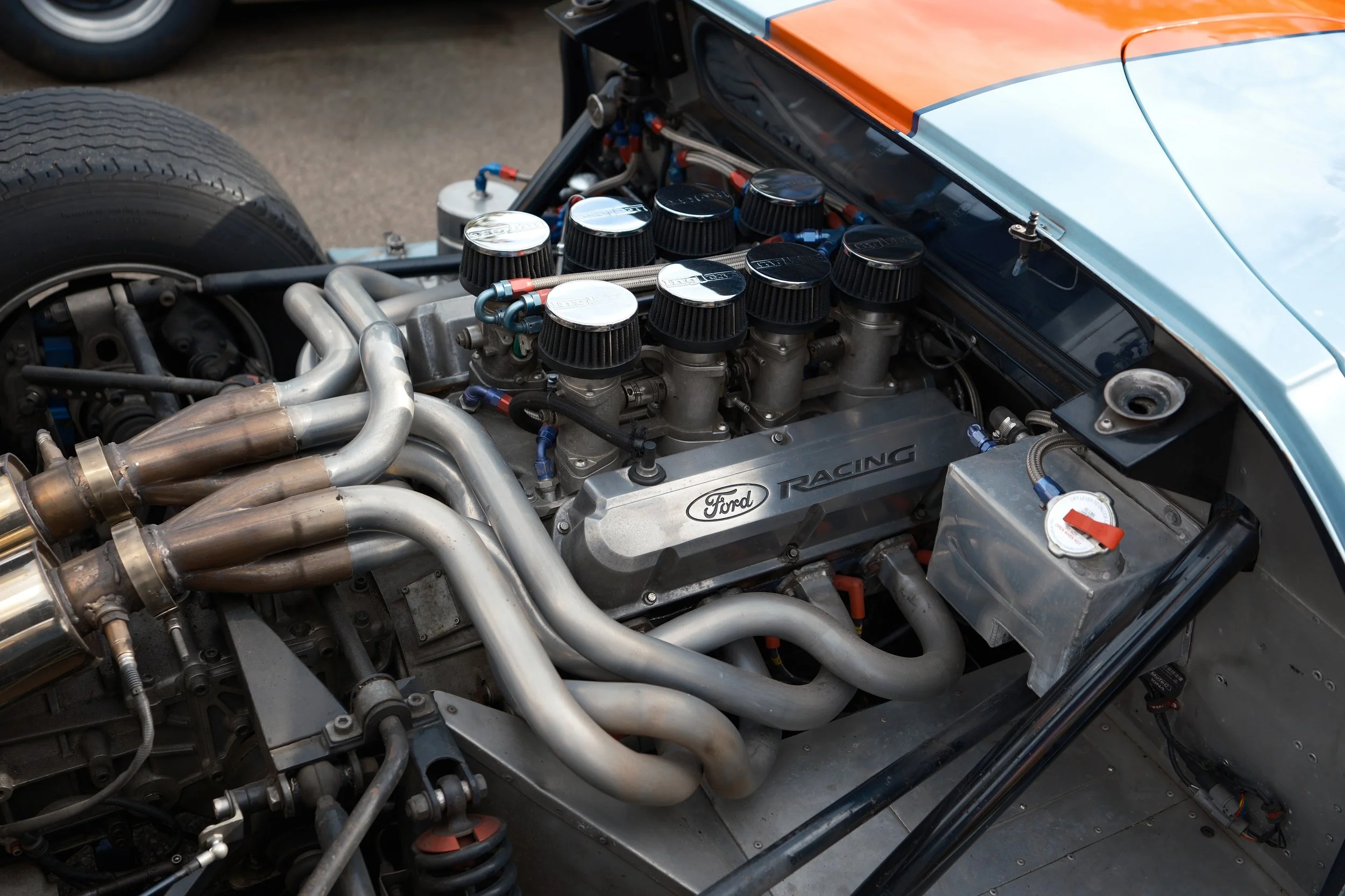 Detailed view of a race car engine with multiple air filters on top, exhaust pipes, hoses, and mechanical components underneath the car’s orange and blue body.