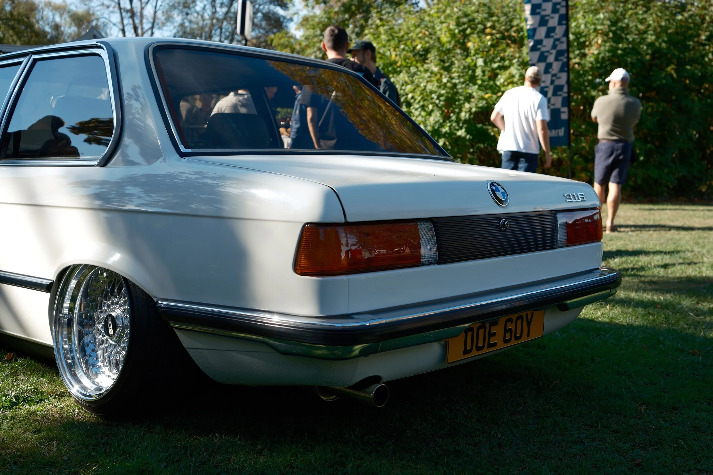 A white vintage BMW 3 Series car parked on grass at an outdoor event with people in the background.