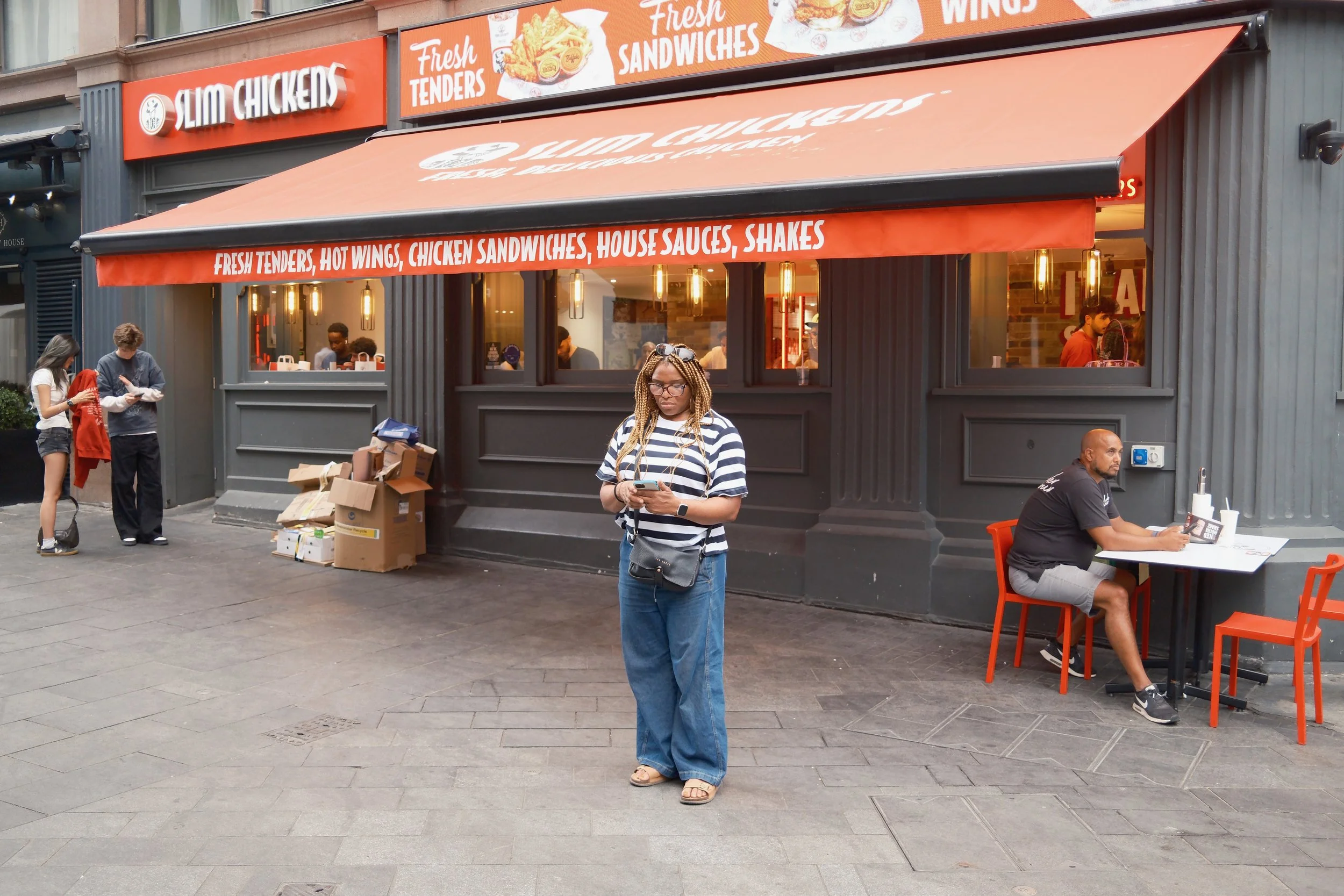 People standing and sitting outside a fast food restaurant called Slim Chickens, which has orange signage and awnings, advertising fresh tenders, hot wings, chicken sandwiches, house sauces, and shakes. A woman with braided hair and glasses, wearing 