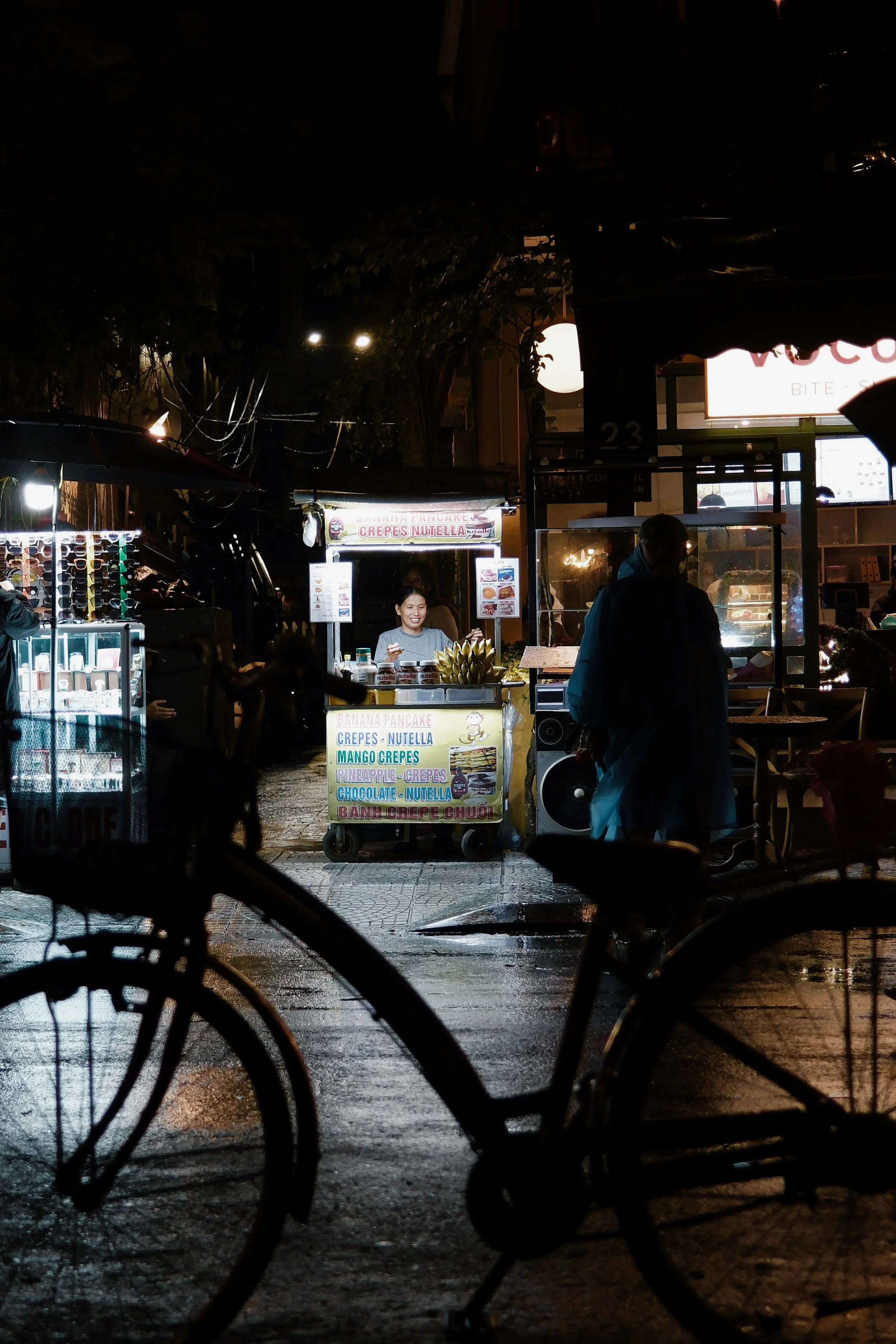 Night scene of street food stall with a smiling woman serving crepes and Nutella, with a colorful menu board listing banana pancakes, mango crepes, pineapple crepes, chocolate Nutella crepes, and bubble tea, seen through silhouetted bicycle wheels in