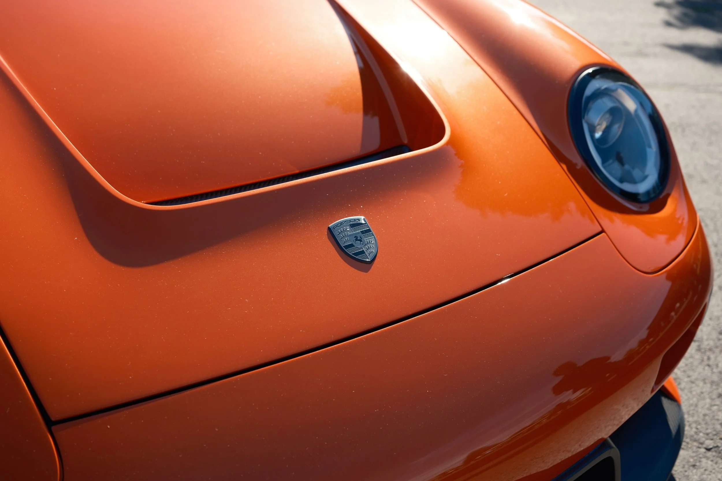 Close-up of an orange sports car hood with a Porsche badge, featuring a prominent hood scoop and a headlight on the right side.