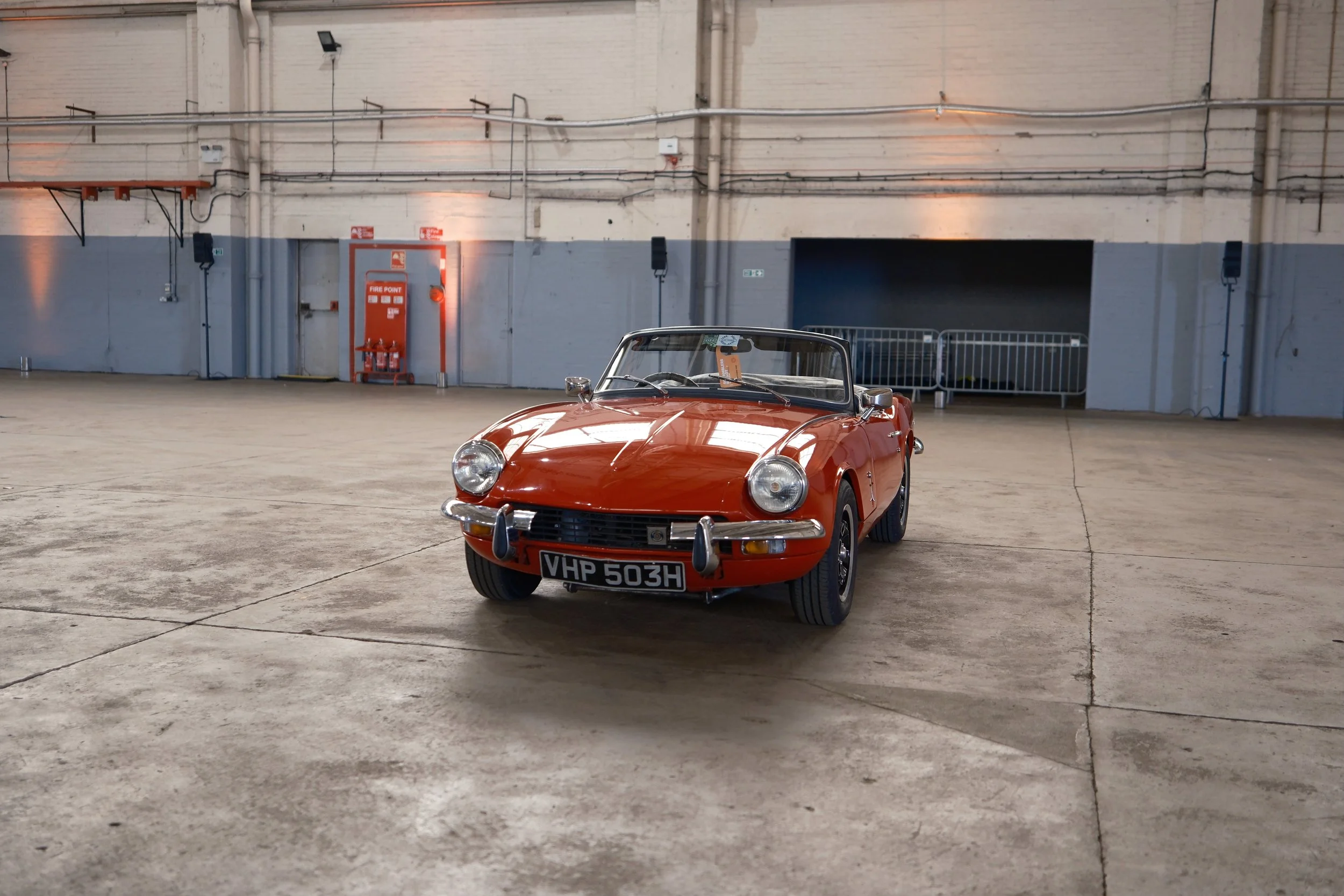 A vintage orange convertible car with a British license plate parked inside a large industrial warehouse with a concrete floor and white brick walls.