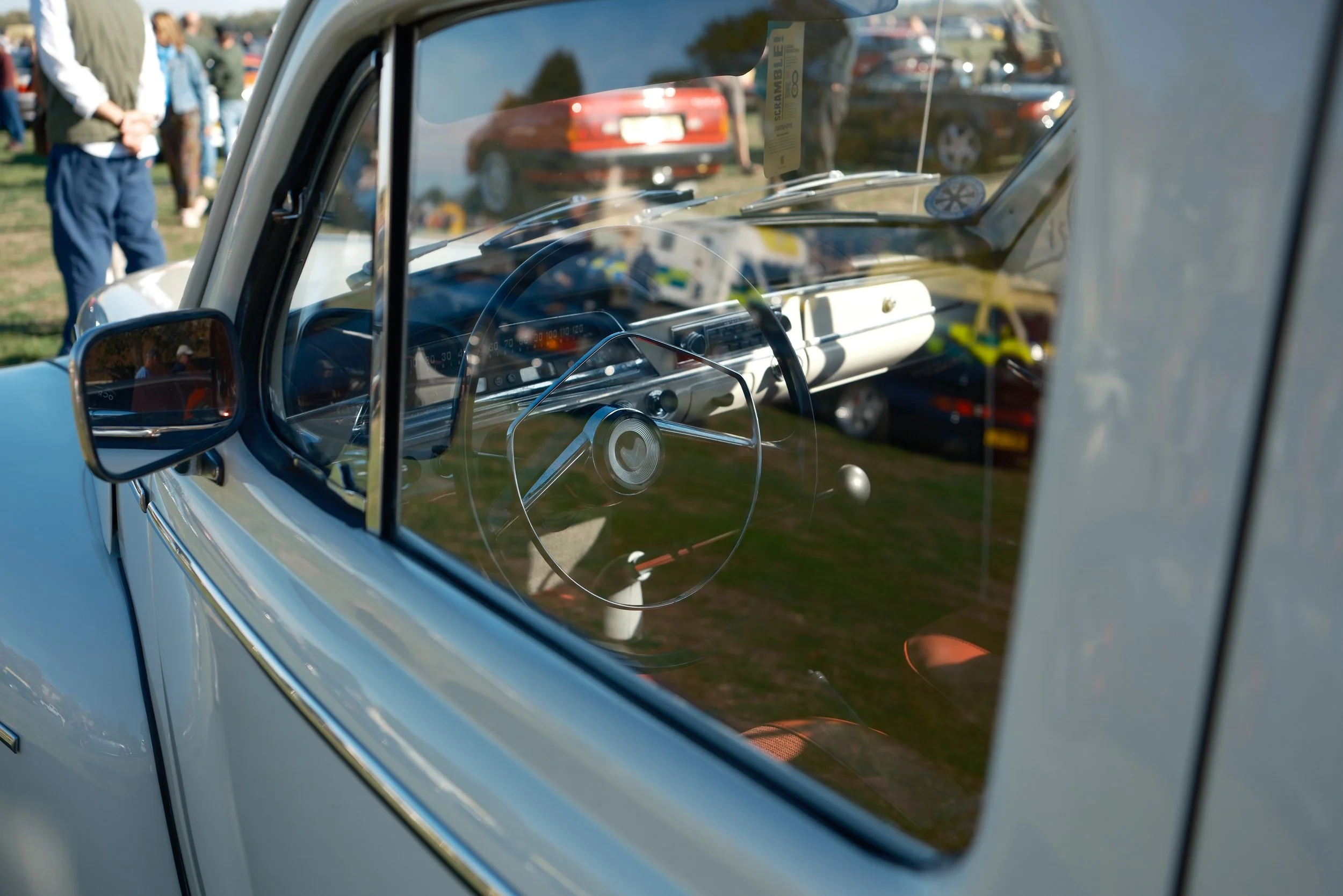 The interior of a vintage car with a view through the window showing a parking lot with multiple cars and people standing around outdoors.