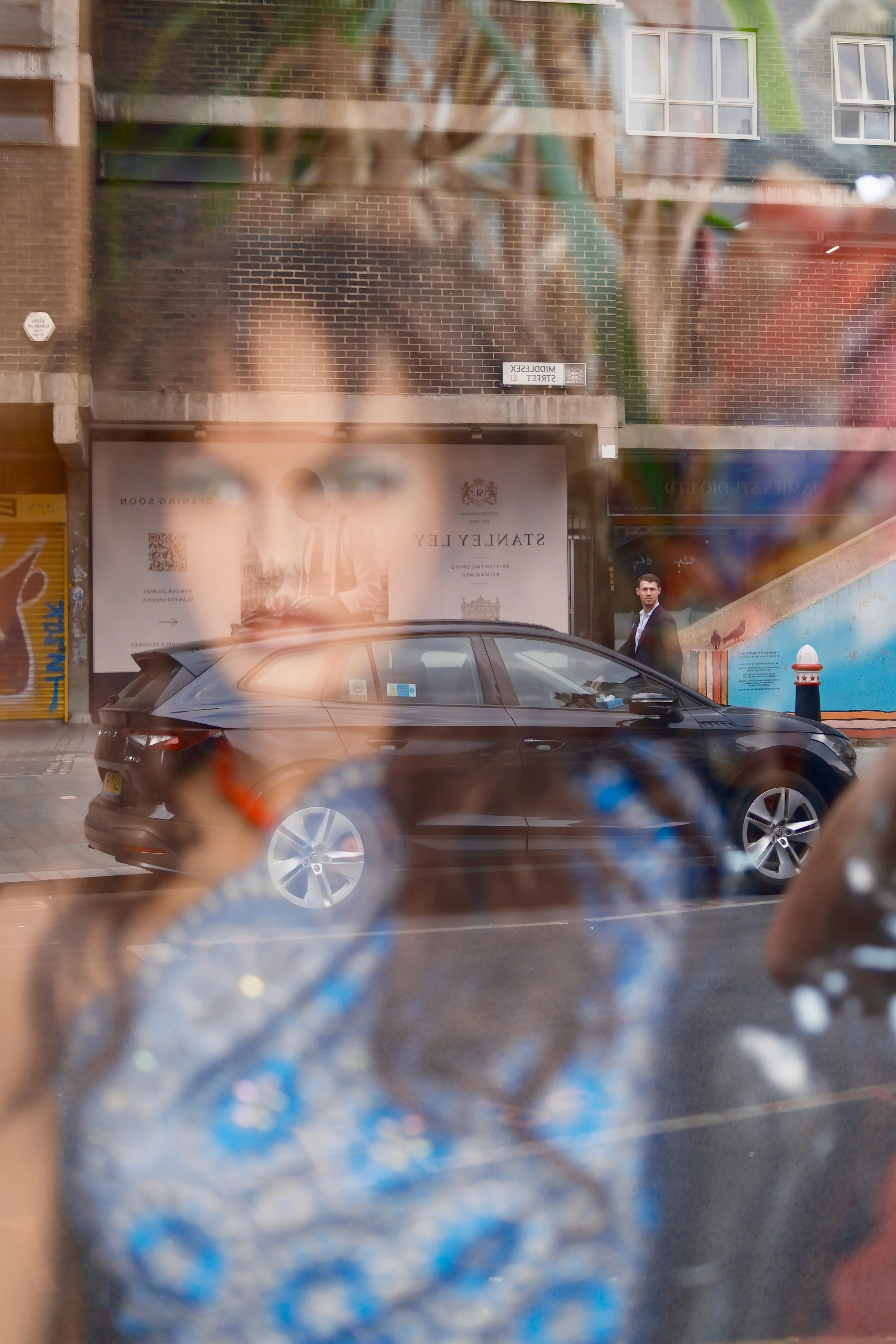 A reflection of a street scene seen through a rain-covered window, including a parked black car, a man in a suit, a brick building with large windows, and a billboard with text.
