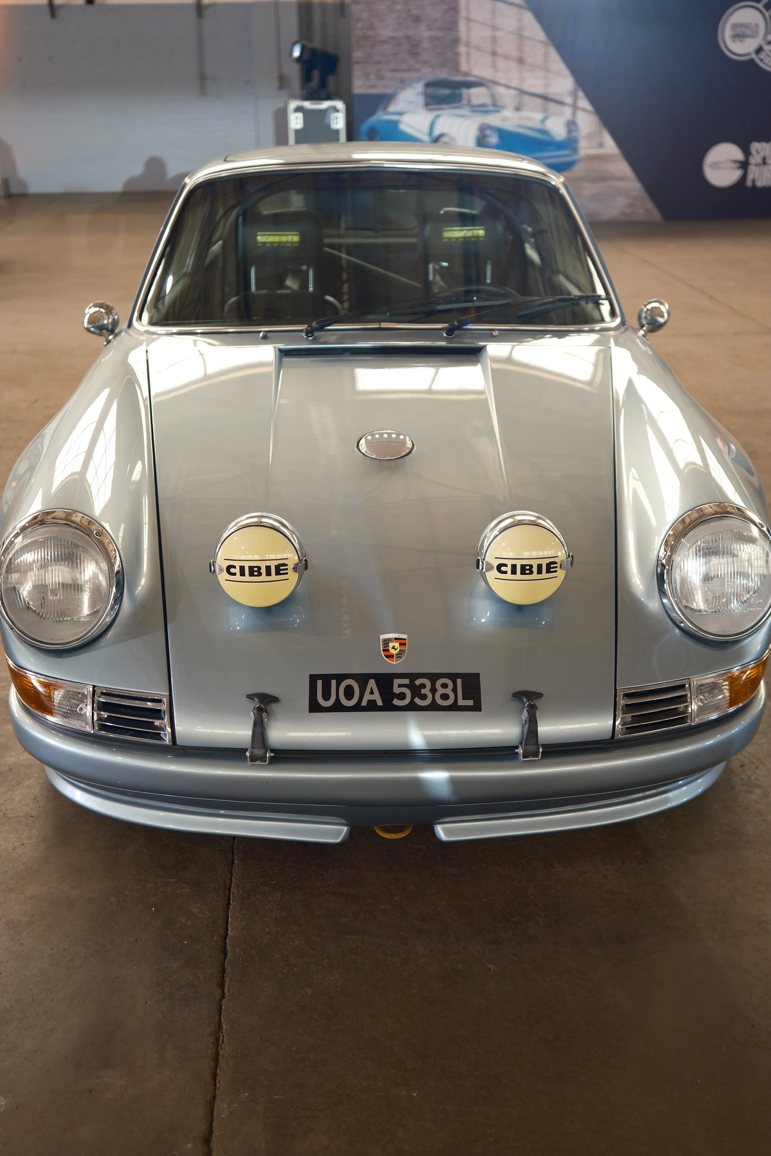 Front view of a vintage silver Porsche 911 race car displayed indoors, with two yellow Cibie rally lights on the front bumper, a license plate reading 'UOA 538L', and a background image of a similar classic car.