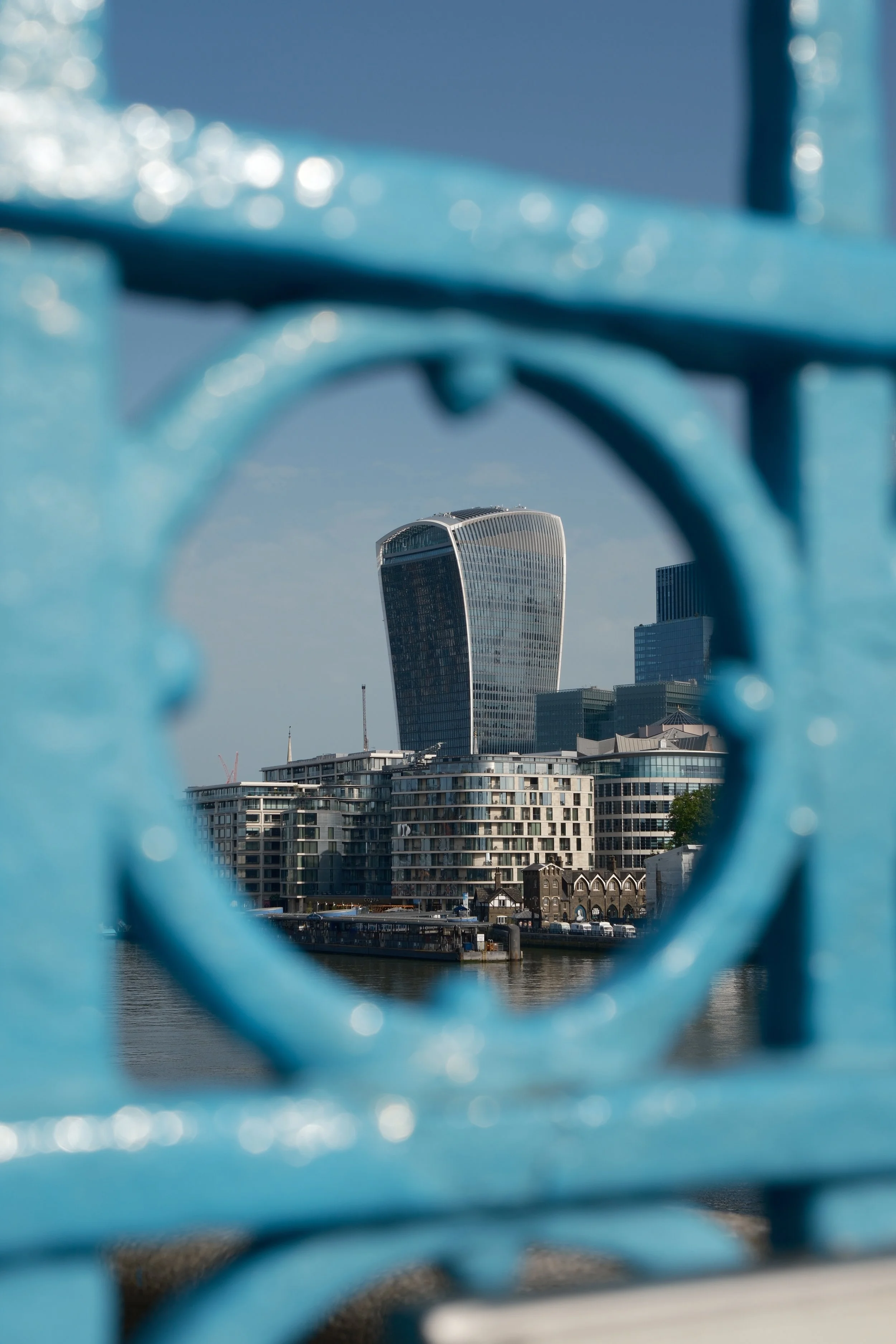 View of a city skyline through a blue-painted metal fence with circular cutouts, showcasing modern skyscrapers and waterfront buildings.