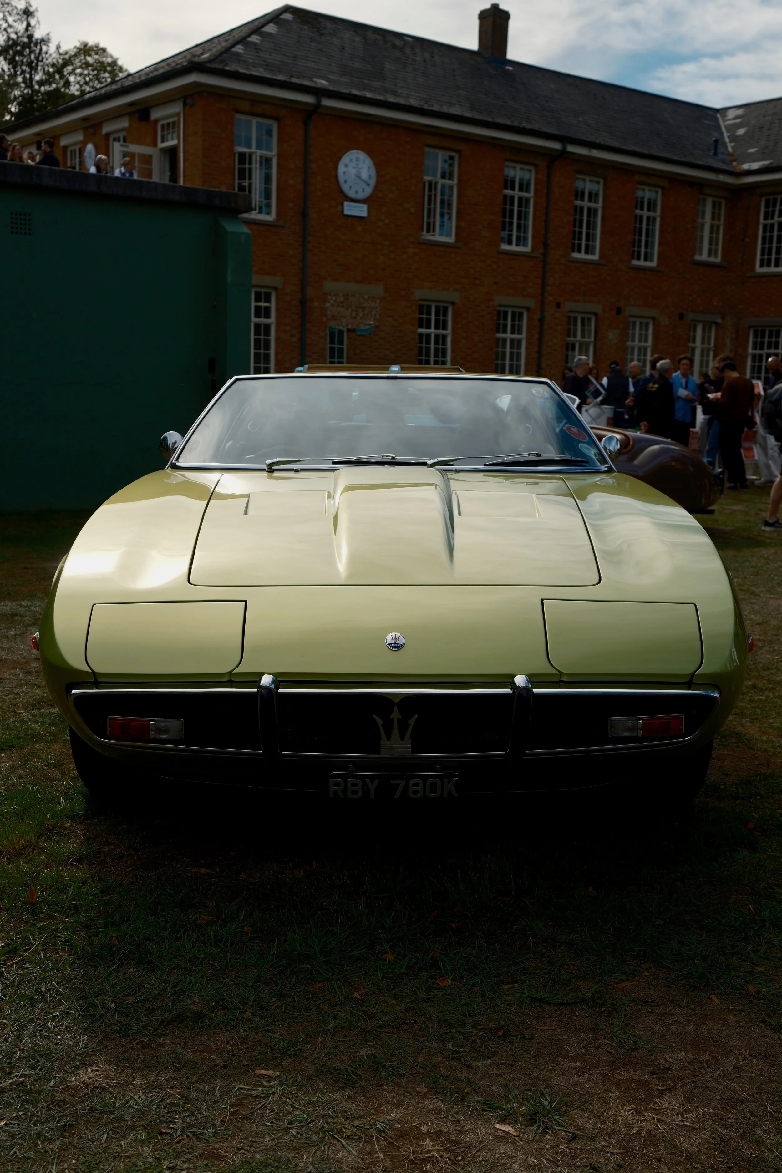 Front view of a vintage yellow sports car parked on grass in front of a brick building with a clock and group of people nearby.