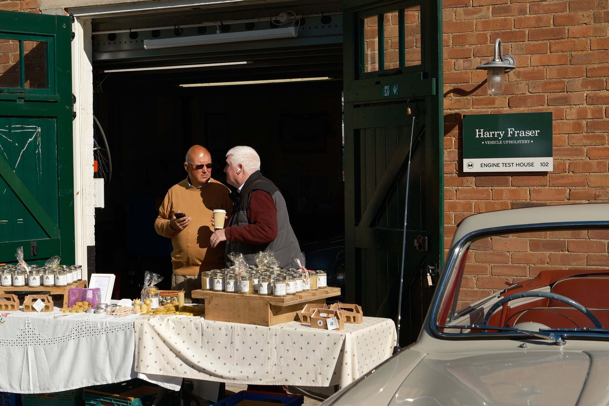 Two men are standing and talking in front of a table filled with small jars, possibly of scented candles or cosmetics, outside a brick building with a sign that reads "Harry Fraser Vehicle Upholstery, Engine Test House 102." There is a display table 