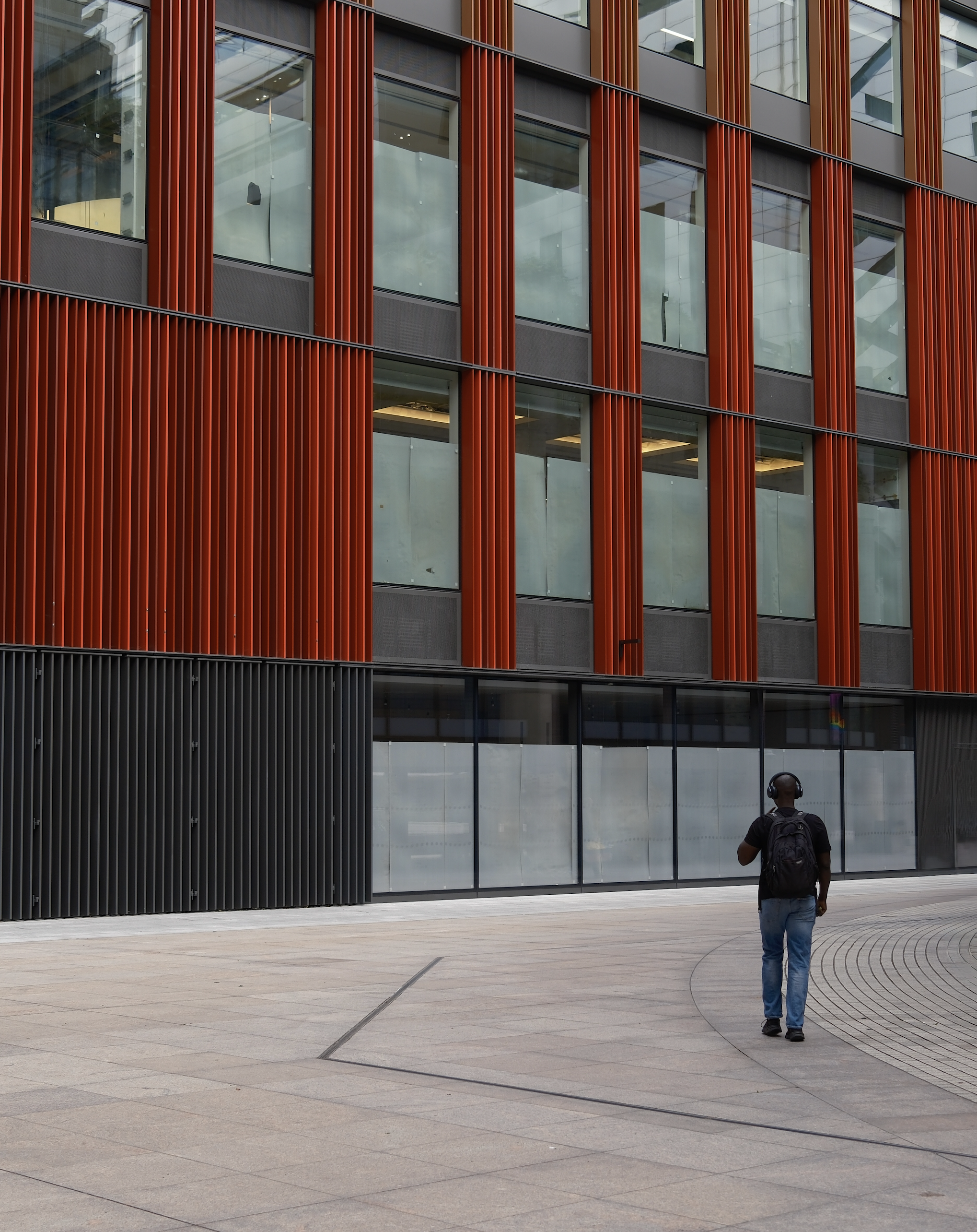 A man with a backpack, wearing headphones, walking in front of a modern building with glass windows and vertical red and black metal panels.