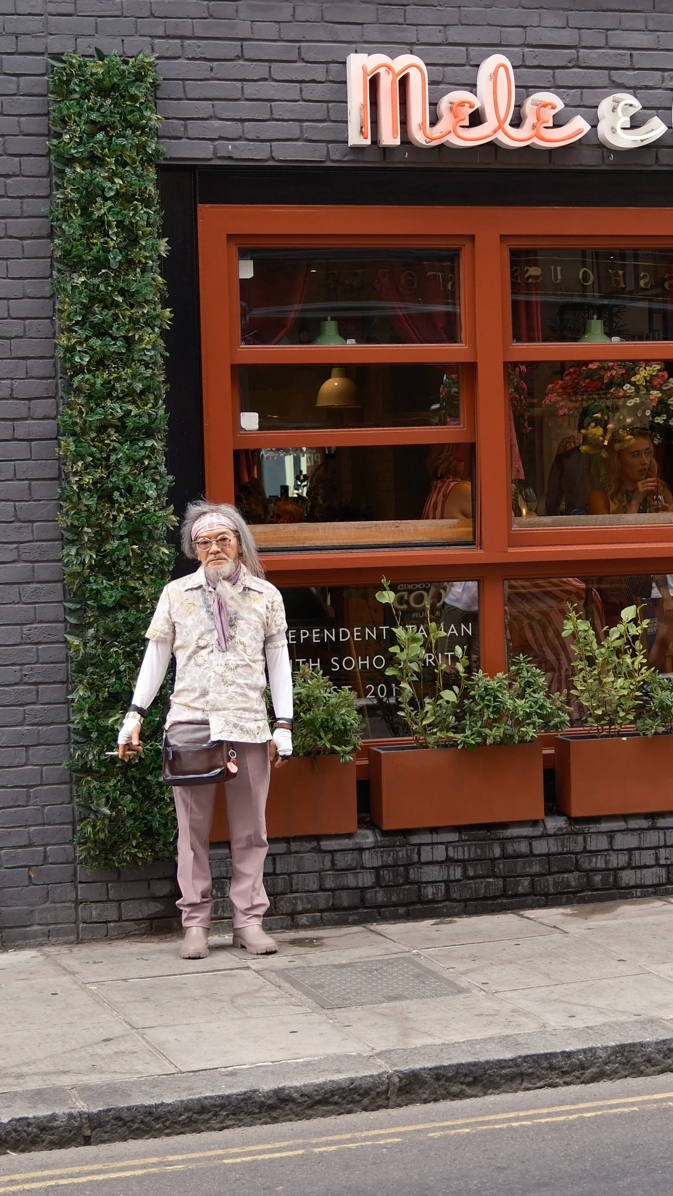 Older man with white hair, glasses, and a beard standing on a city sidewalk outside a cafe with a brick and greenery exterior, holding a purse and wearing a light-colored shirt, pants, and boots.