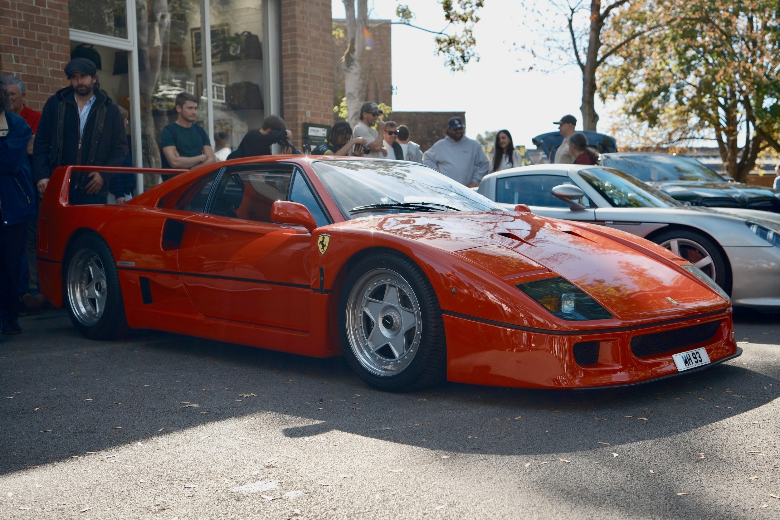 Red Ferrari sports car parked with a crowd of people standing behind it during a car show on a sunny day with autumn trees in the background.