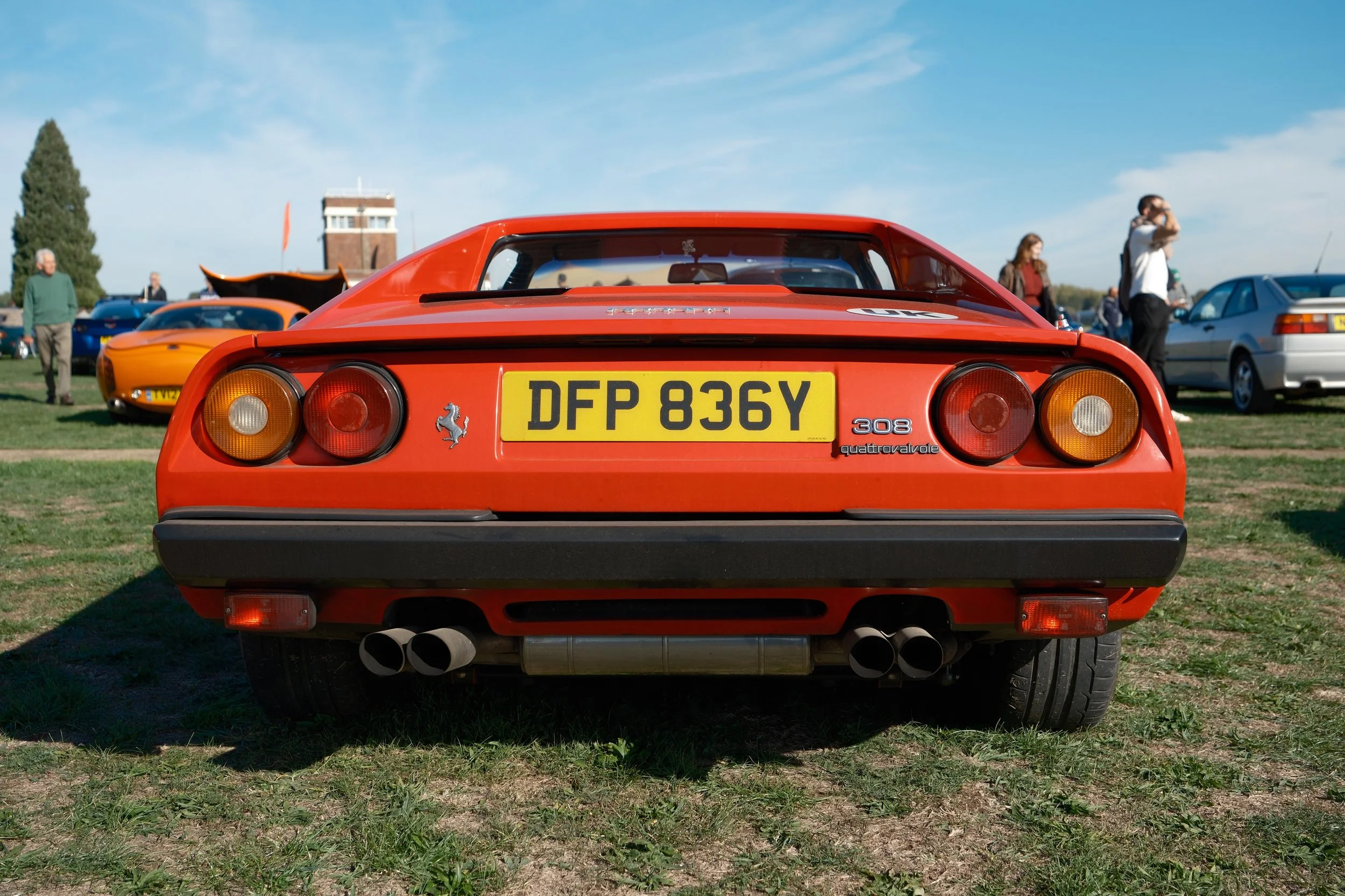 Rear view of a red Ferrari 308 with a yellow license plate reading DFP 836Y at a car show, with other cars and people in the background under a clear blue sky.