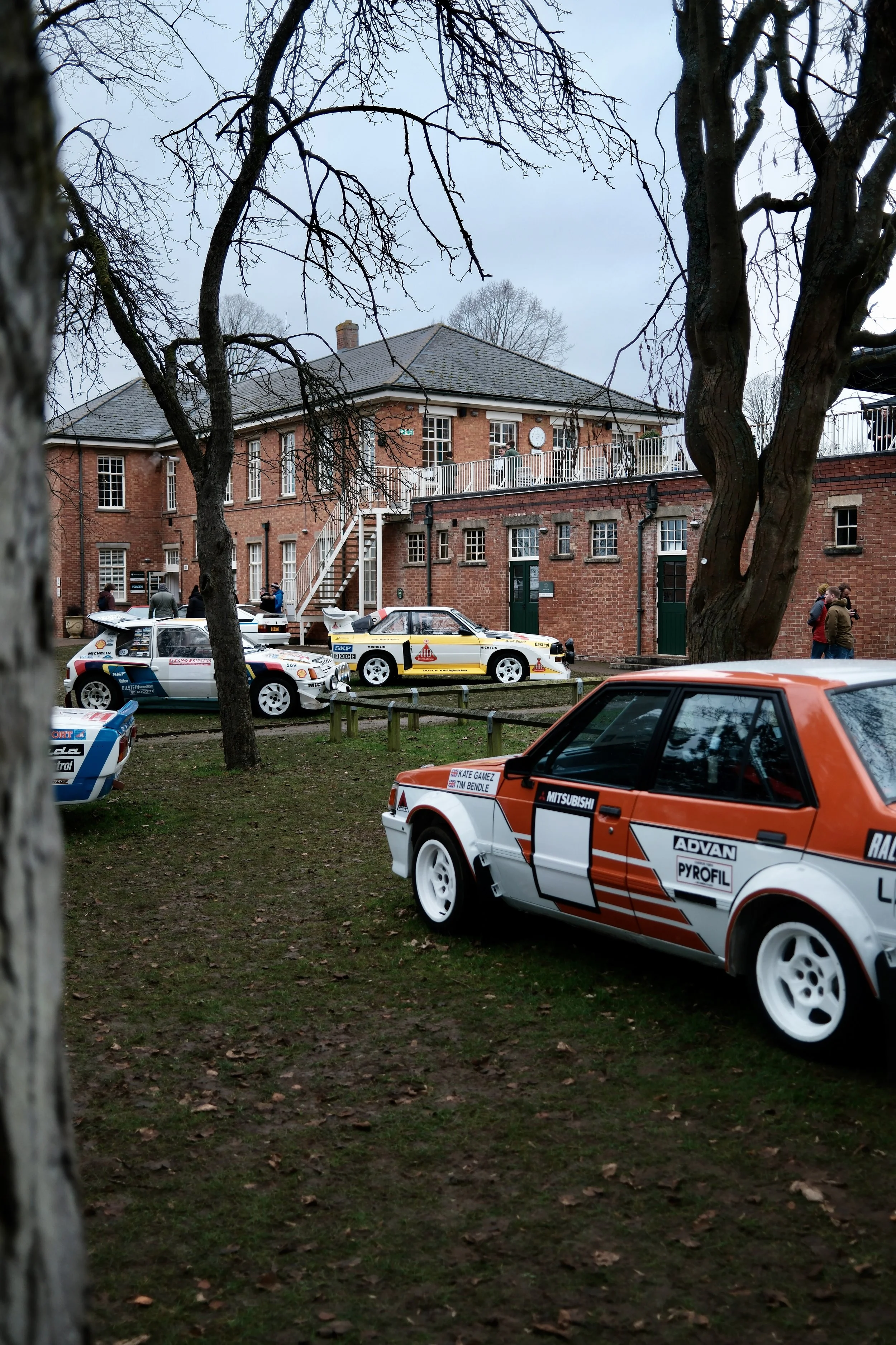 A collection of rally cars parked on grass in front of a brick building with trees and people around.