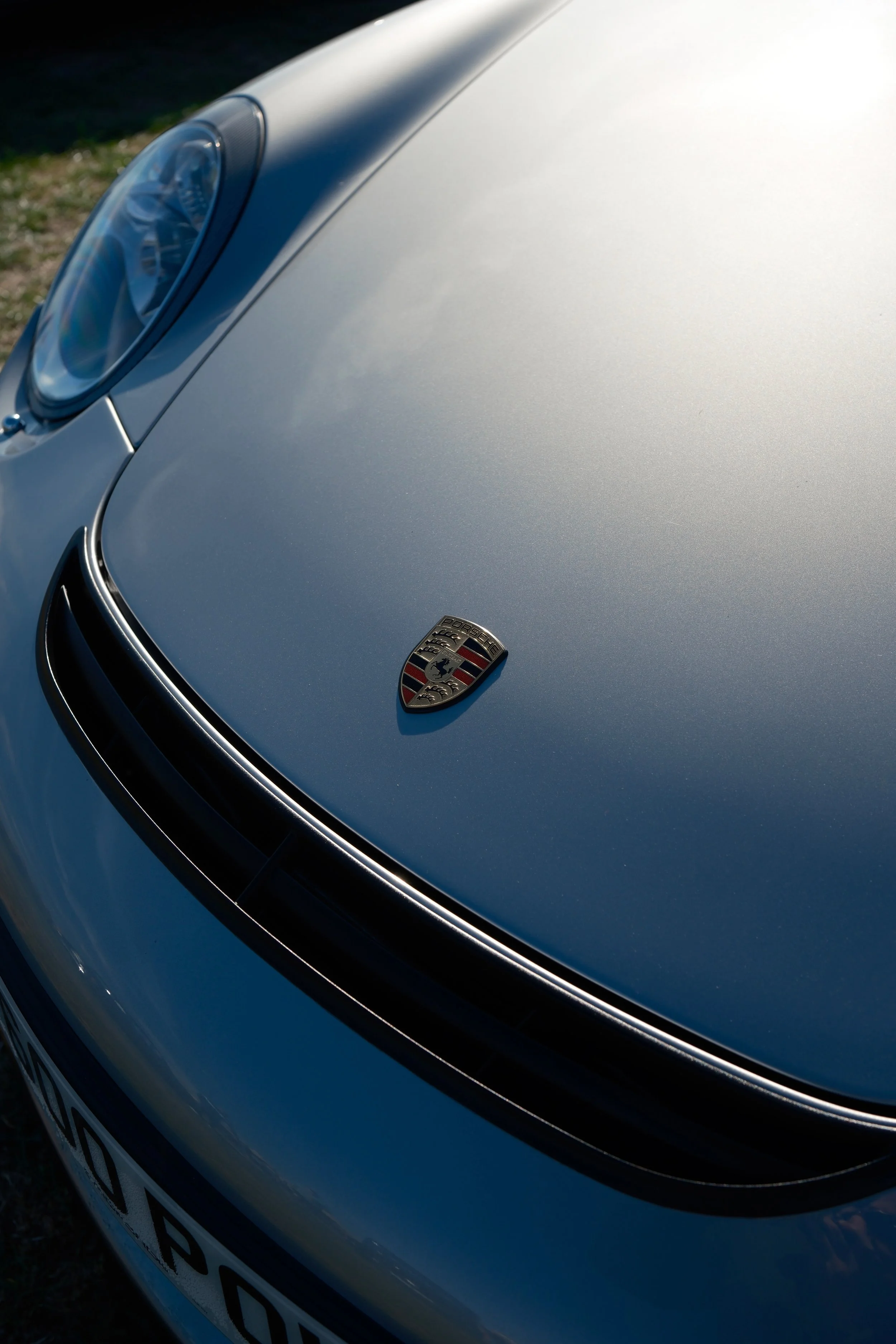 Close-up of the front of a silver Porsche car, showing the Porsche emblem on the hood and part of the headlight.