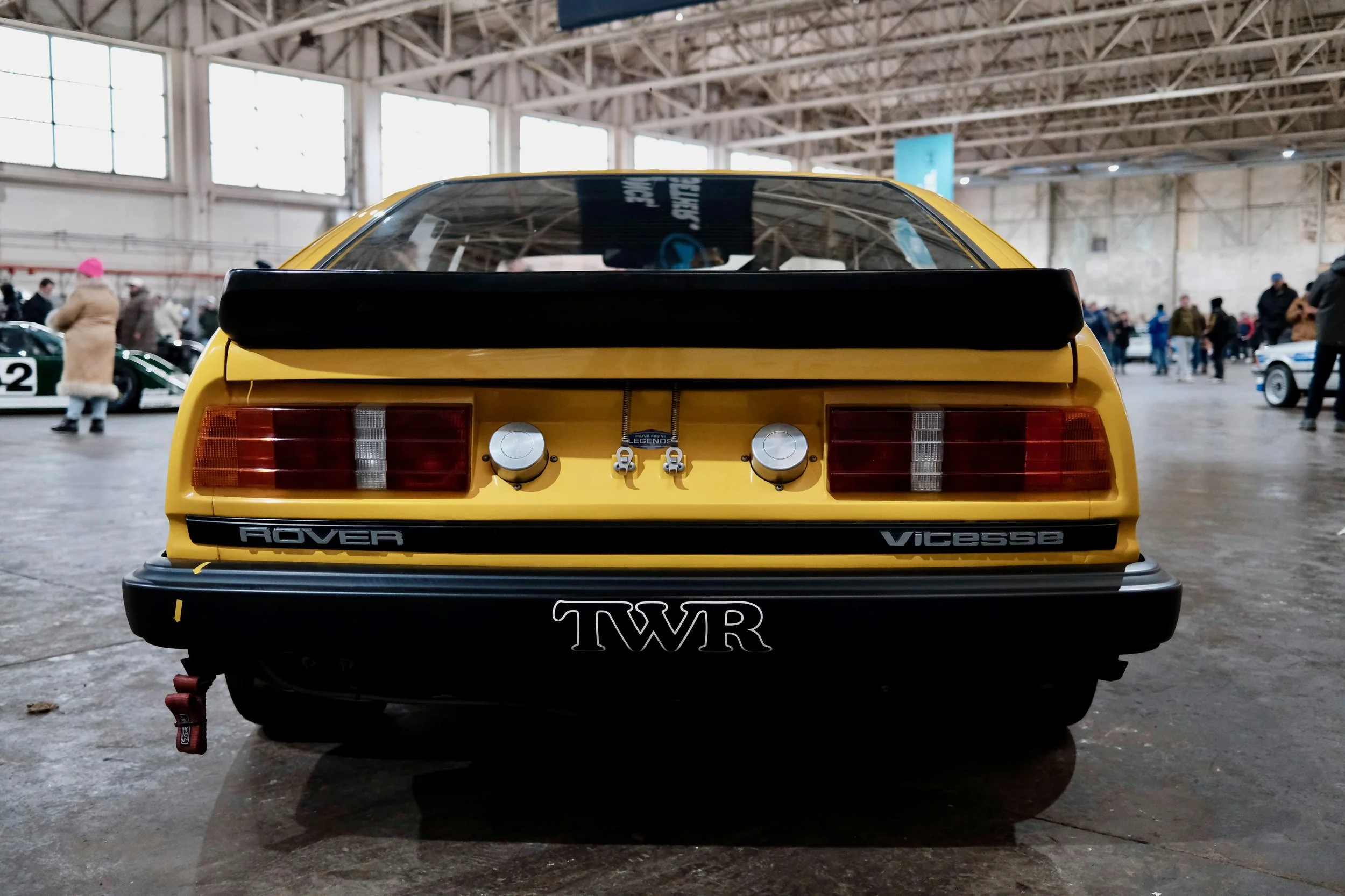 Rear view of a yellow classic sports car in an indoor exhibition with people walking around, large windows, and a high ceiling.