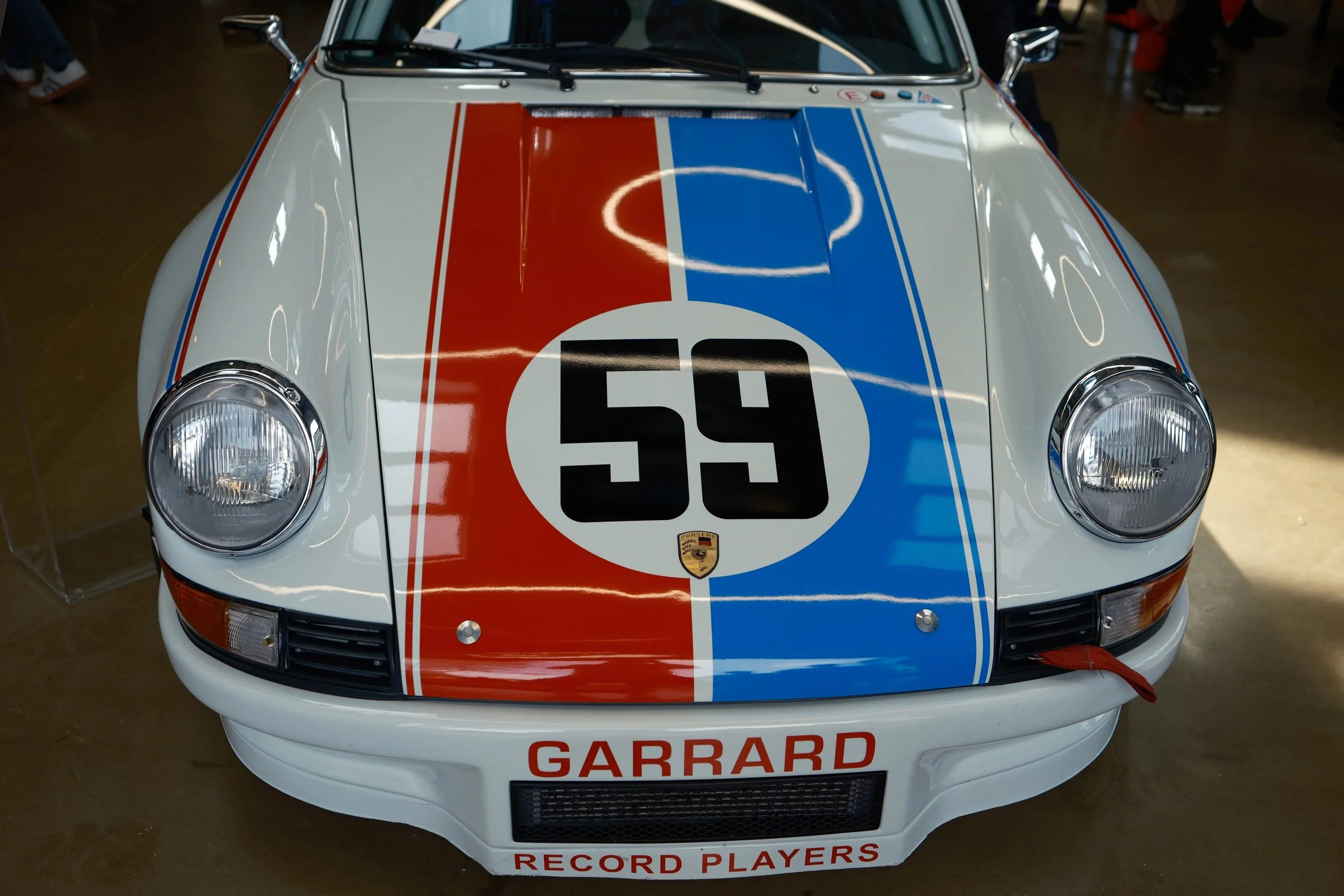Front view of a vintage Porsche race car with a white body featuring a red and blue racing stripe design, a large number 54 inside a circular background, the Porsche logo, and the words "GARRARD" and "RECORD PLAYERS" on the front bumper.