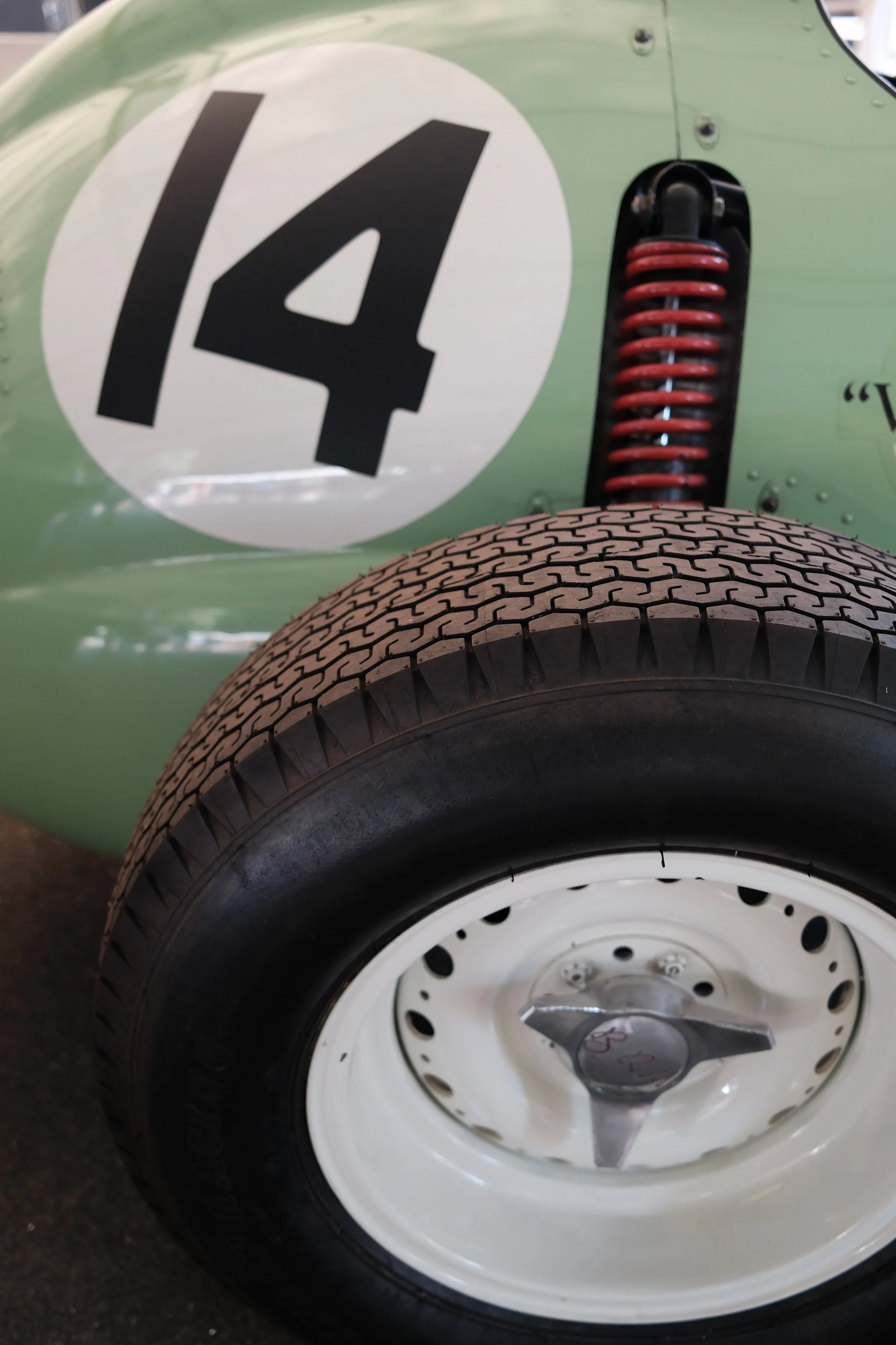 Close-up of a green vintage race car with a white circle logo displaying the number 4, a red suspension coil, and a black tire with a white rim.