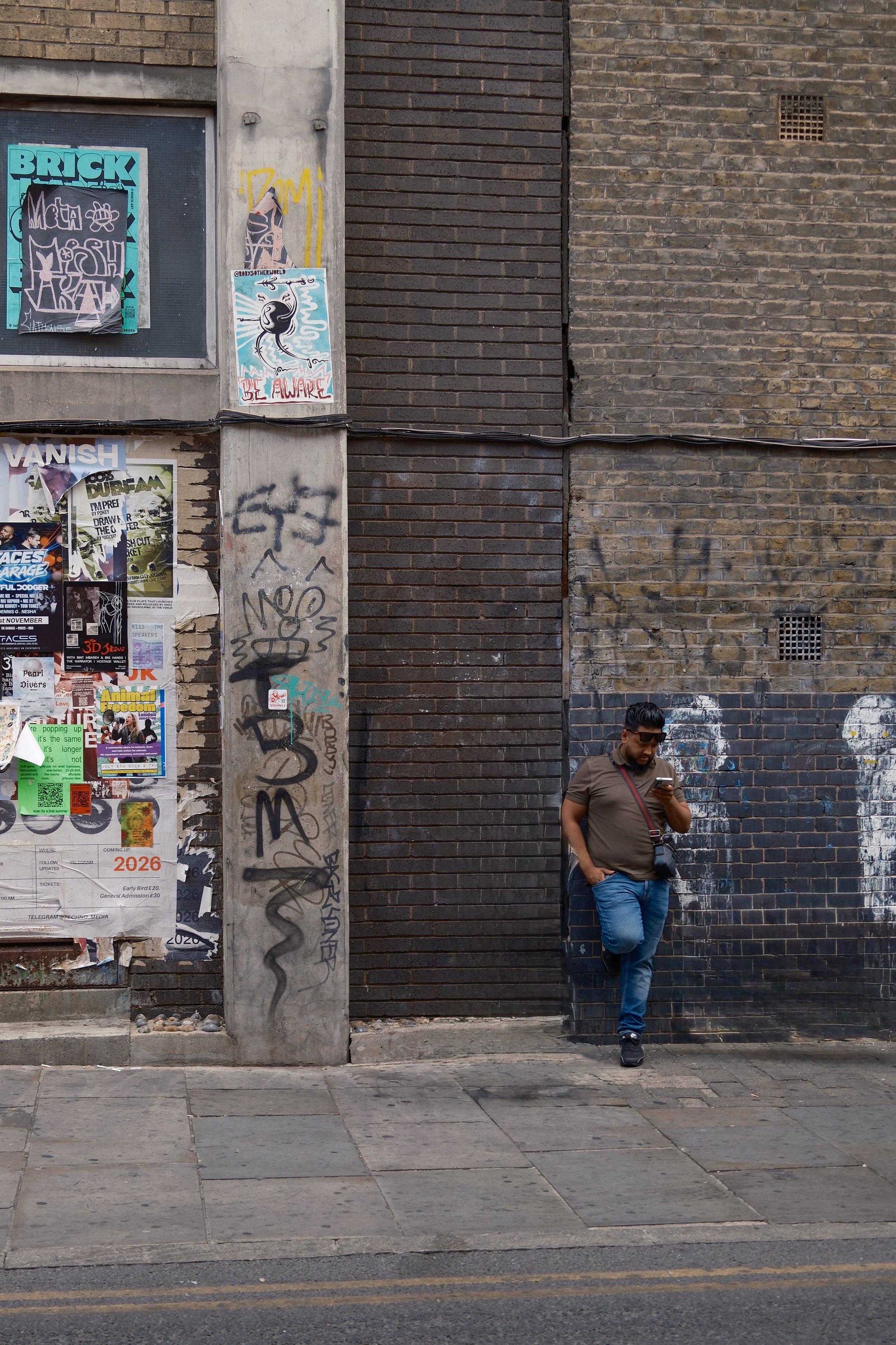 A man wearing sunglasses, a brown shirt, and jeans is standing on a city sidewalk, looking at his phone, in front of a brick wall with graffiti and posters.