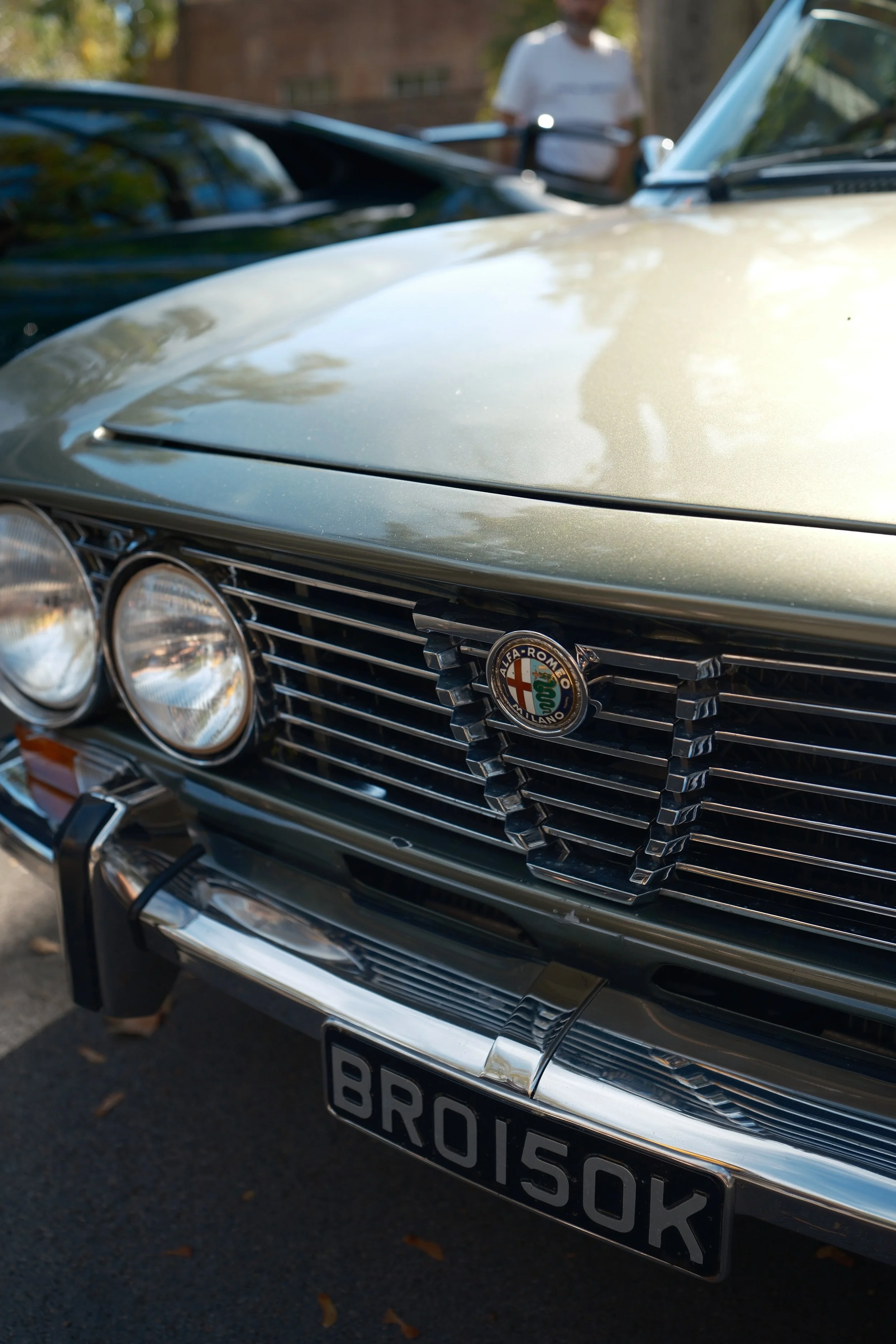 Close-up of a classic black Alfa Romeo car's front grille and headlights, with a partial view of the hood and a European-style license plate.