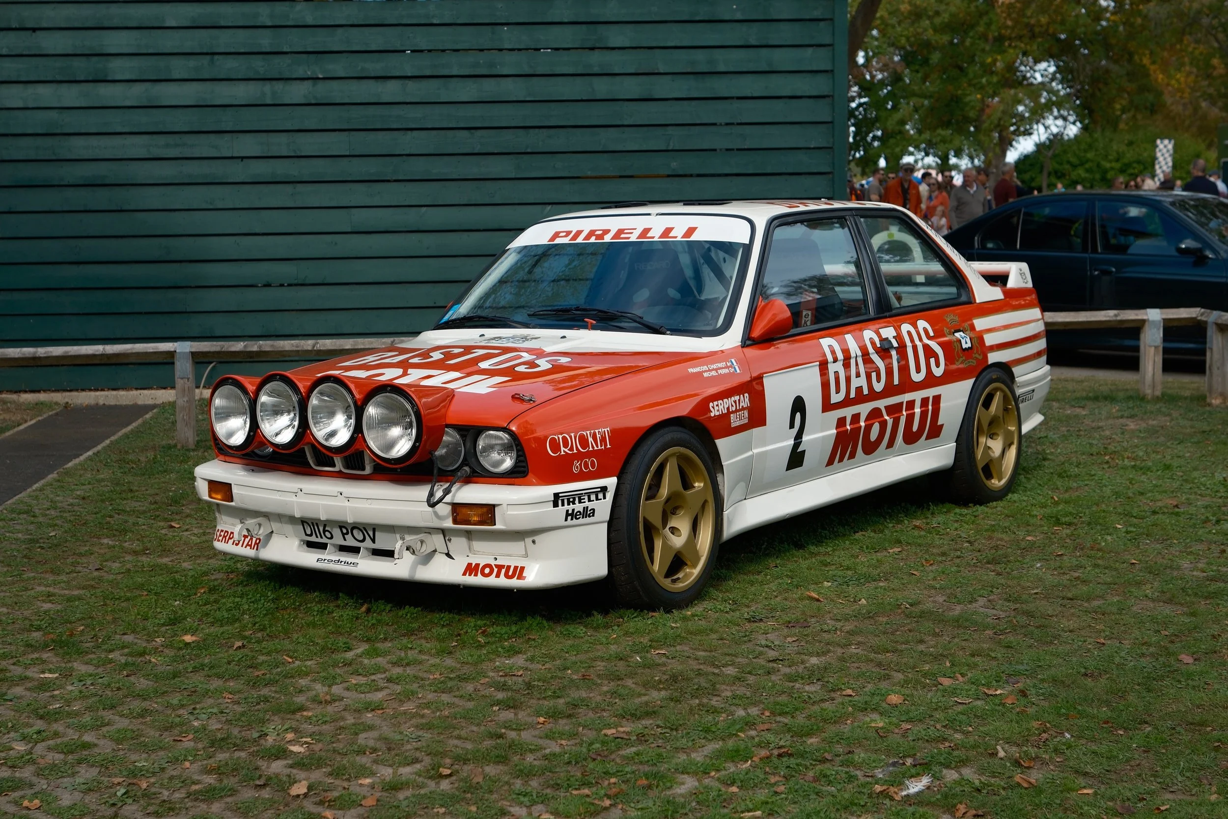 A classic rally car with a red and white racing livery parked on grass, with a group of people and trees in the background.