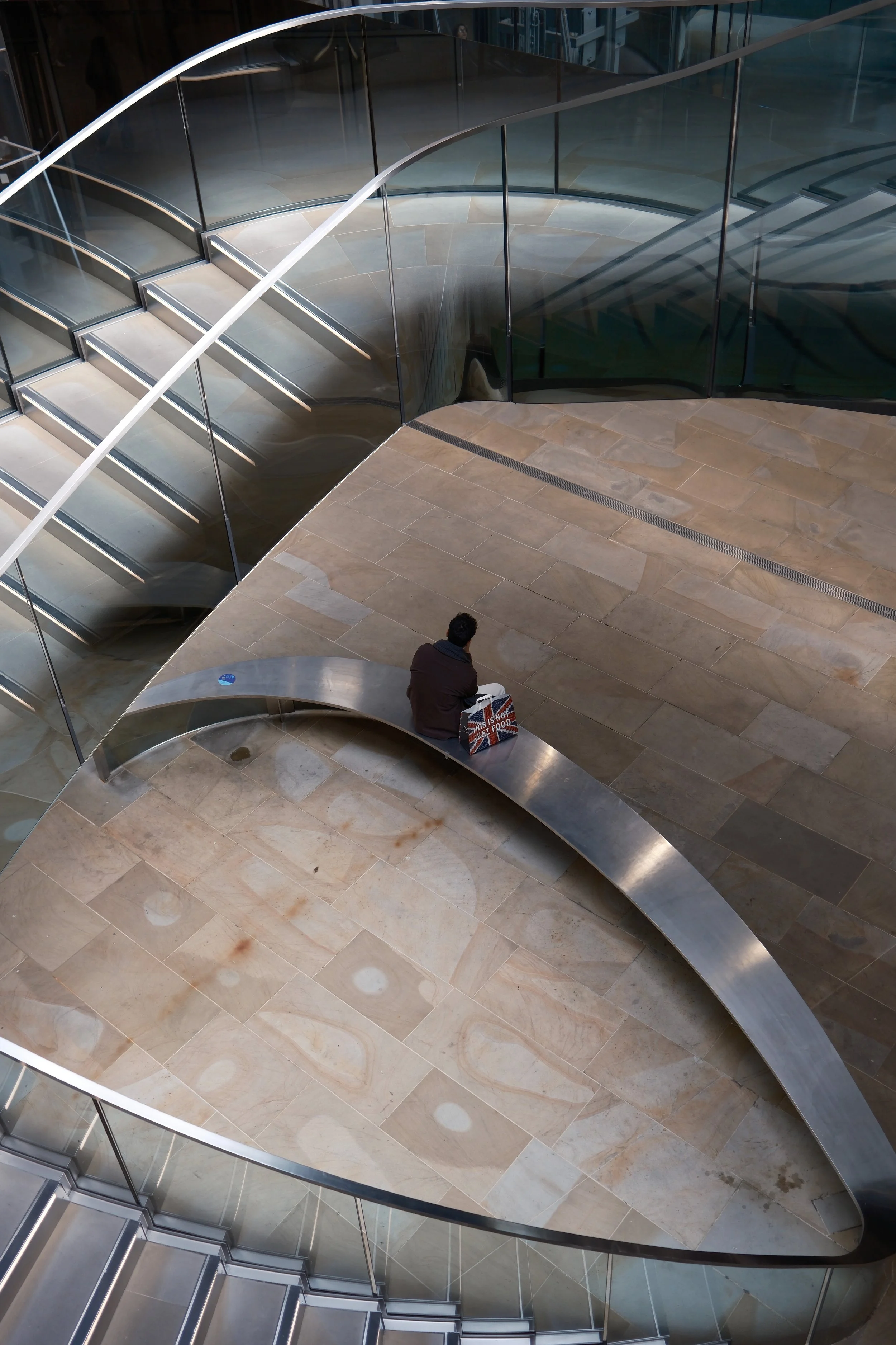 Interior view of a modern building featuring a spiral staircase with metal and glass railings, beige tiled floor, and a person seated on a curved bench holding a shopping bag.