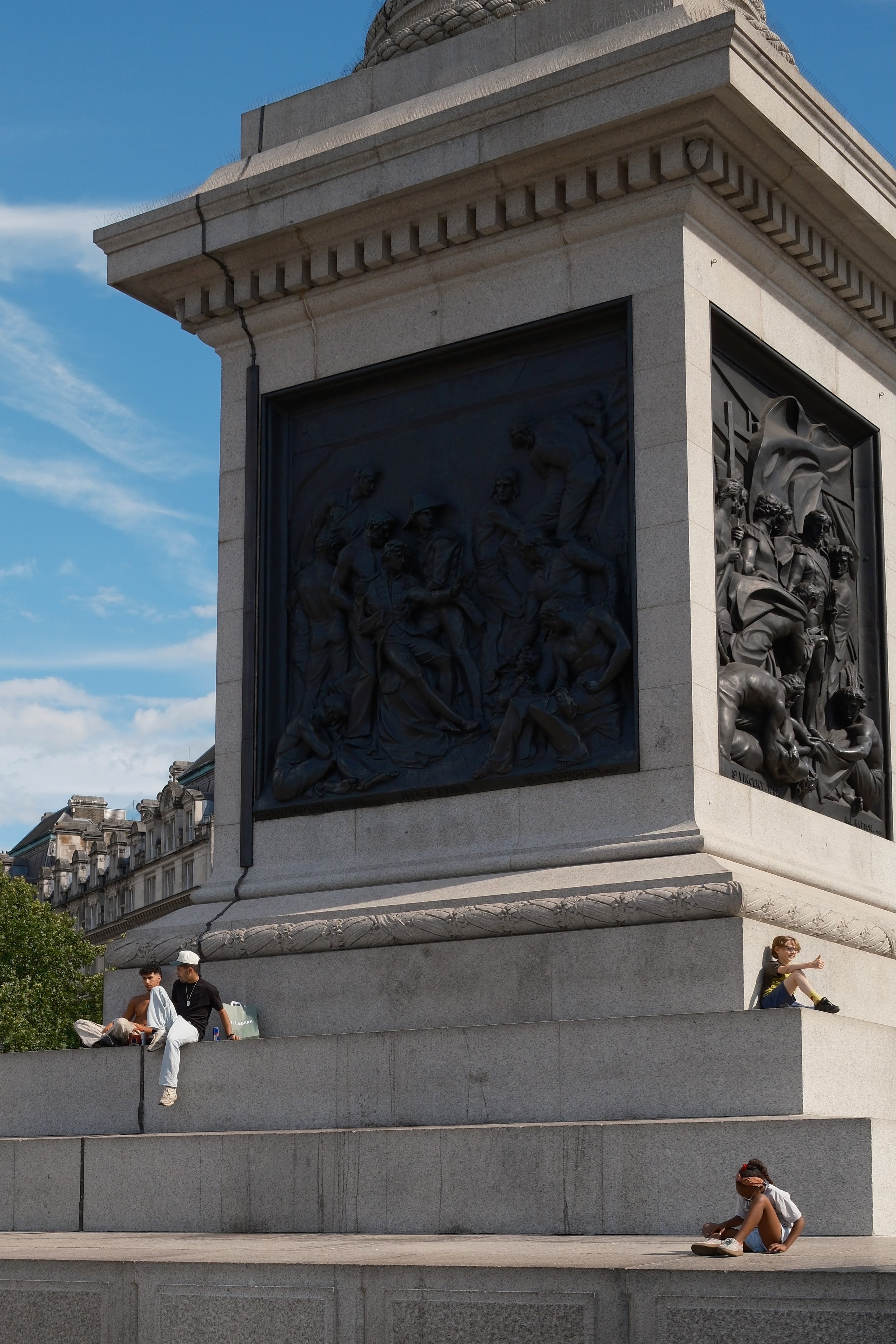 Close-up of a monument or memorial with detailed bas-reliefs of soldiers. People sitting and standing around the monument, including a woman sitting on the lower step and two men sitting on the middle step, one wearing a cap. Blue sky with some cloud