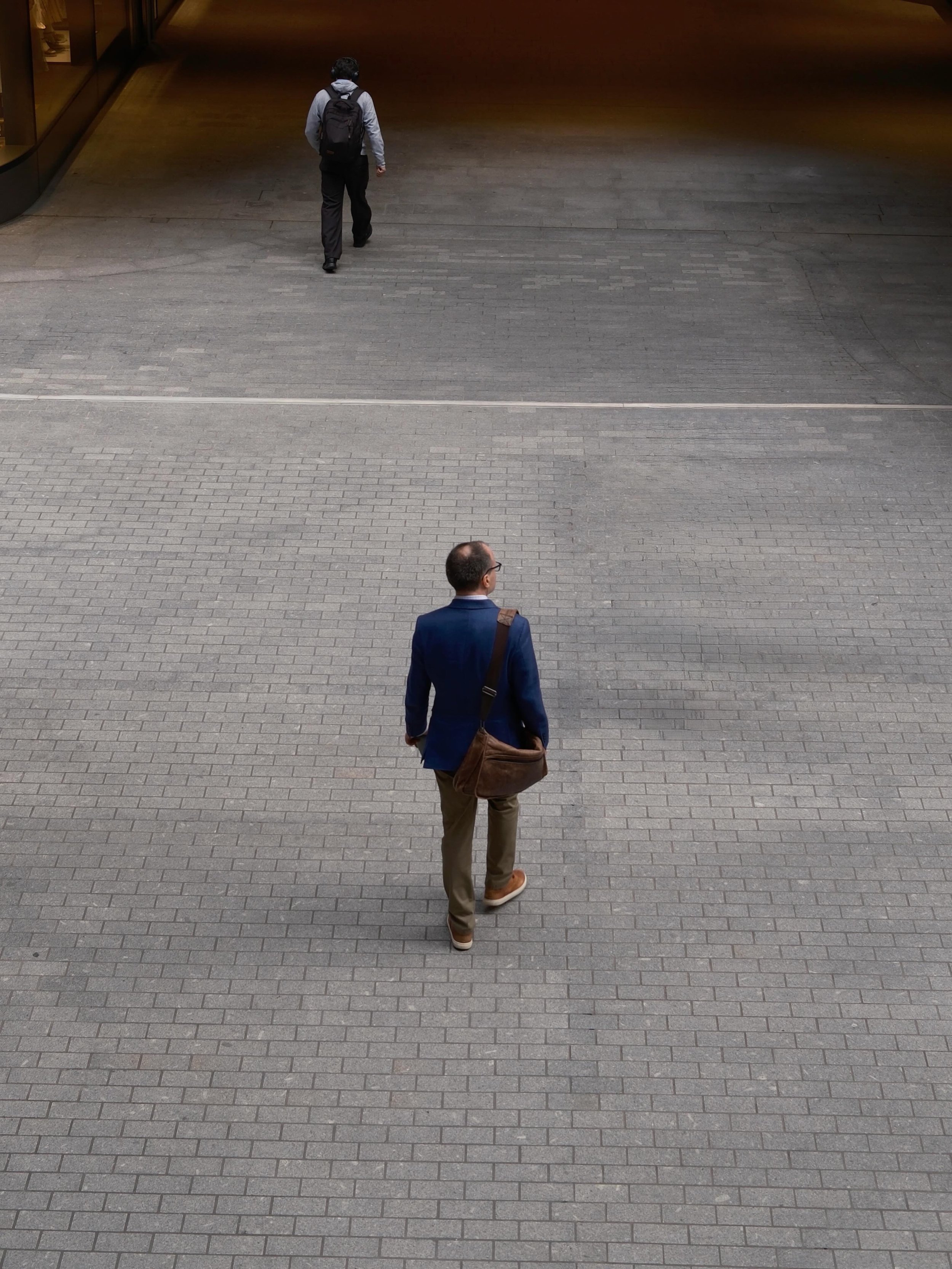 Two men walking on a paved area outside a building. One man, in the foreground, wears a blue blazer, khaki pants, and carries a brown bag. The other man, further back, wears a gray hoodie and black pants, with a backpack, walking away from the camera