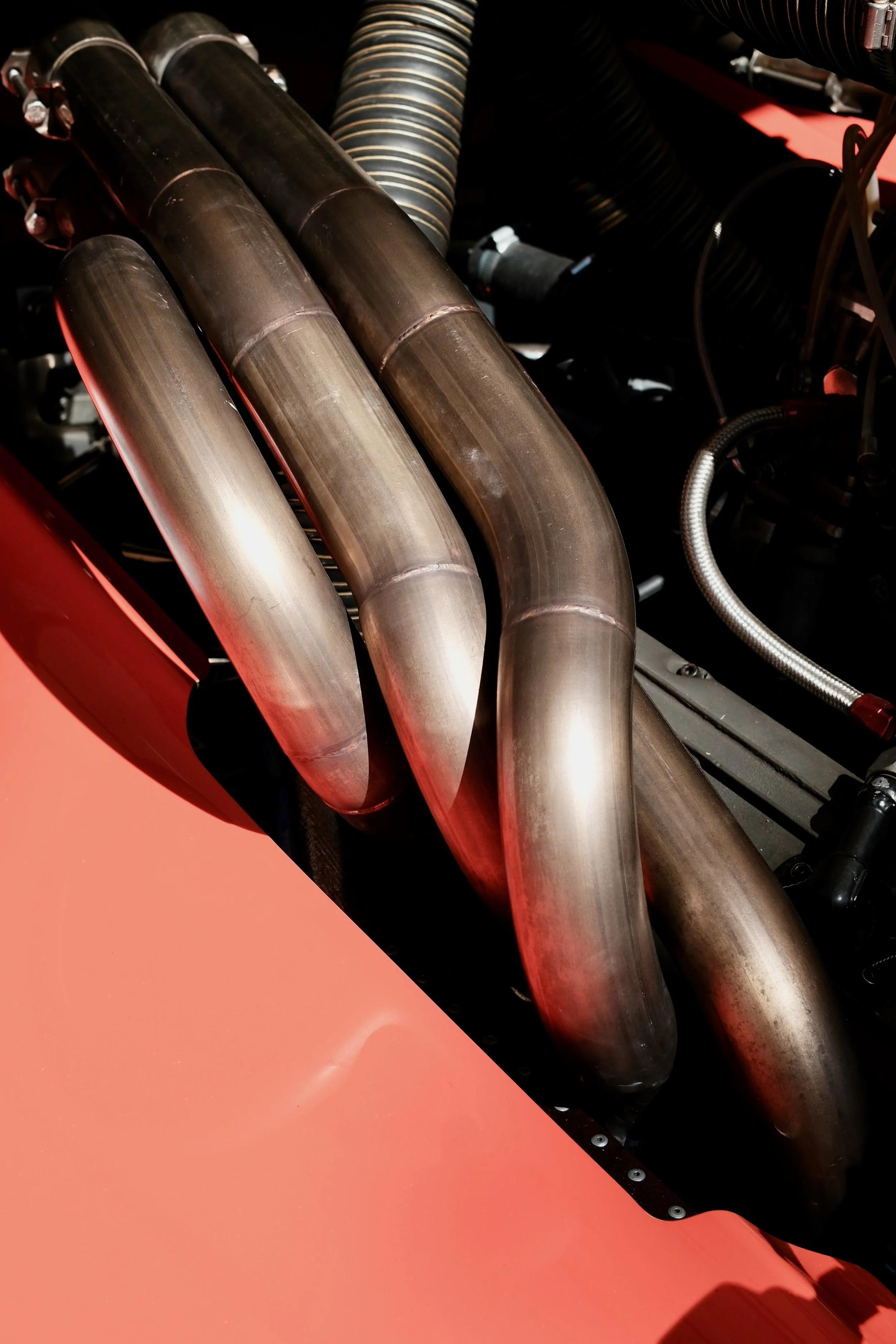 Close-up view of a row of metallic exhaust headers on a high-performance engine, with a black heat shield and red background.
