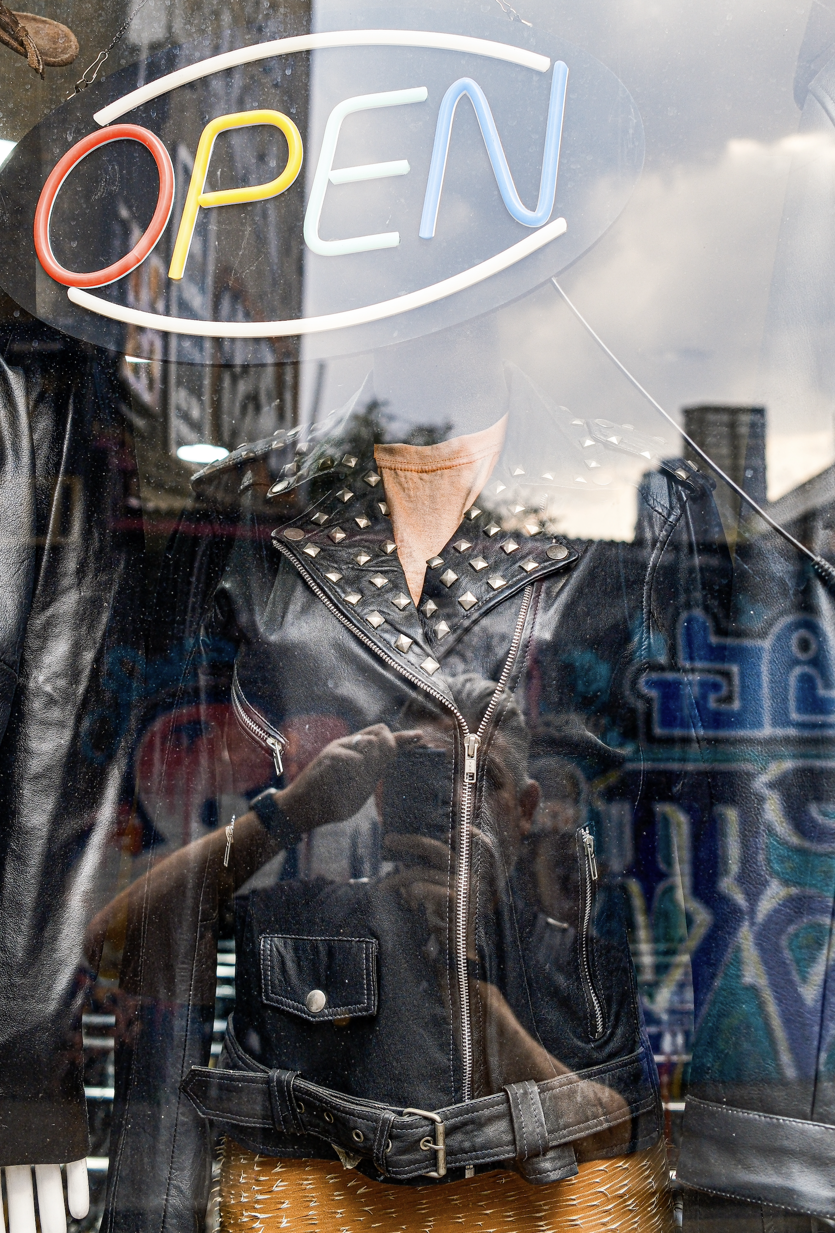 A mannequin dressed in a black leather jacket with silver studs, a pink shirt, and a yellow-patterned skirt, visible through a store window with an illuminated "OPEN" neon sign.