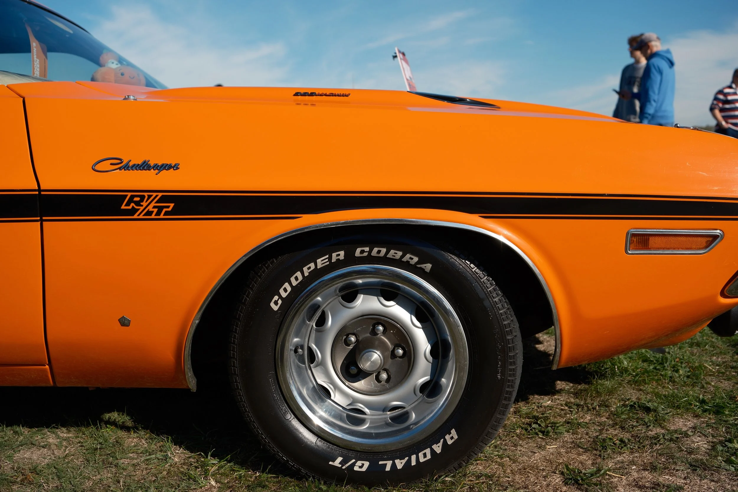 Close-up of an orange Dodge Challenger car, showing the front left side including the wheel, hood, and part of the windshield with reflection of a toy figure. Three people are in the background, standing and looking at their phones.