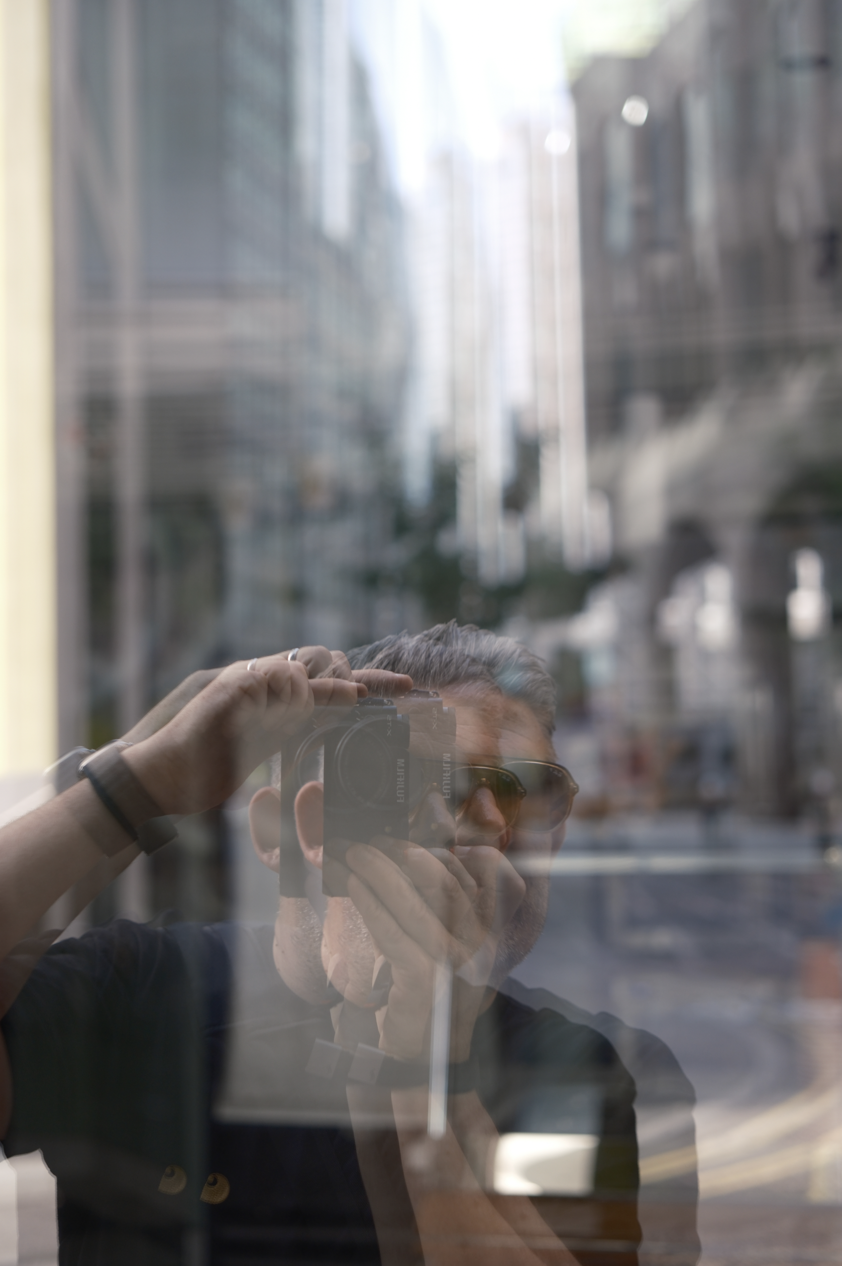 Reflection of a man wearing sunglasses taking a photo with a camera, seen through a glass window with a blurry urban background.