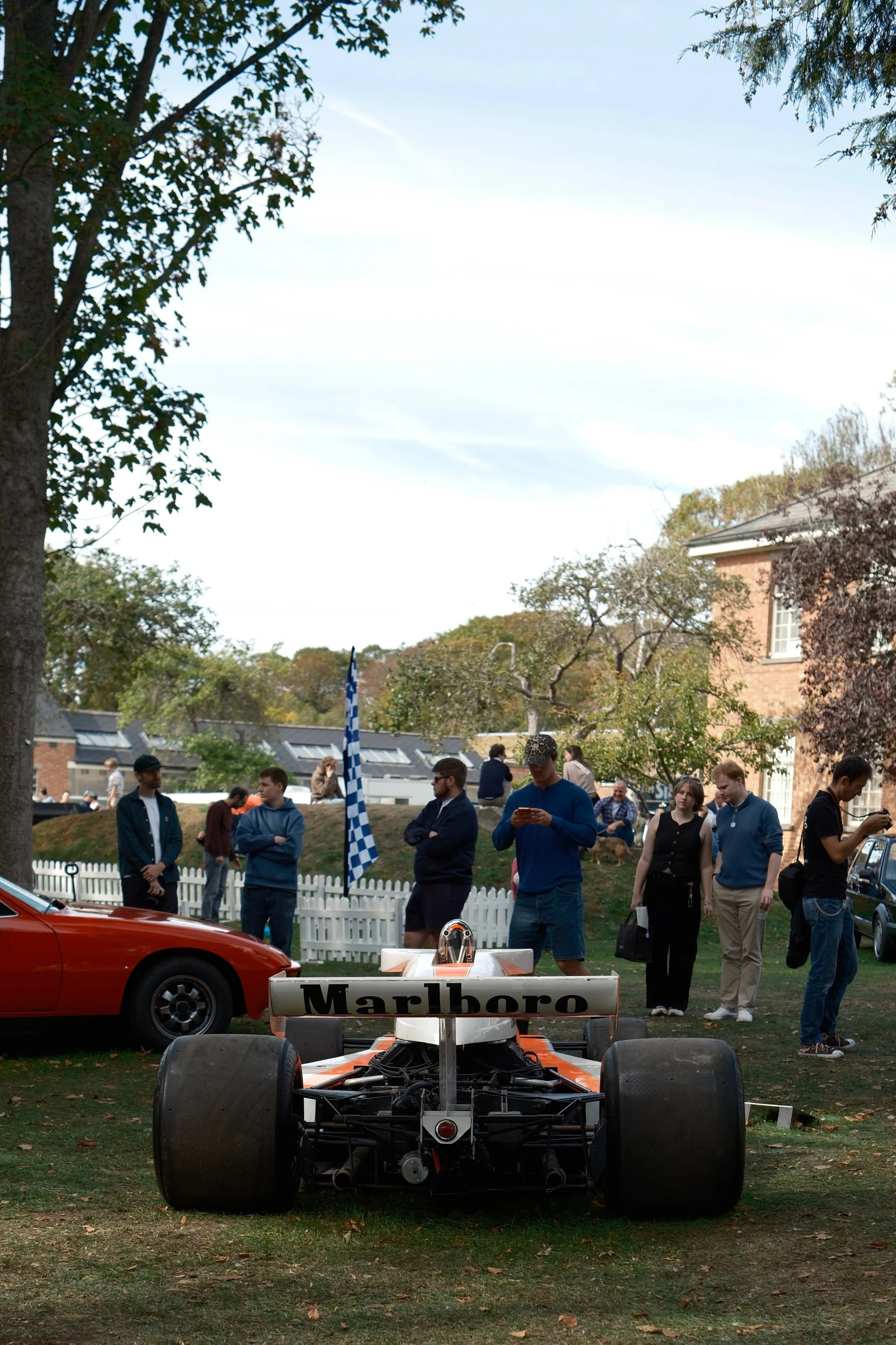 A crowd of people gathered outdoors at a car and race car display event, with trees, a house, and a white picket fence in the background.