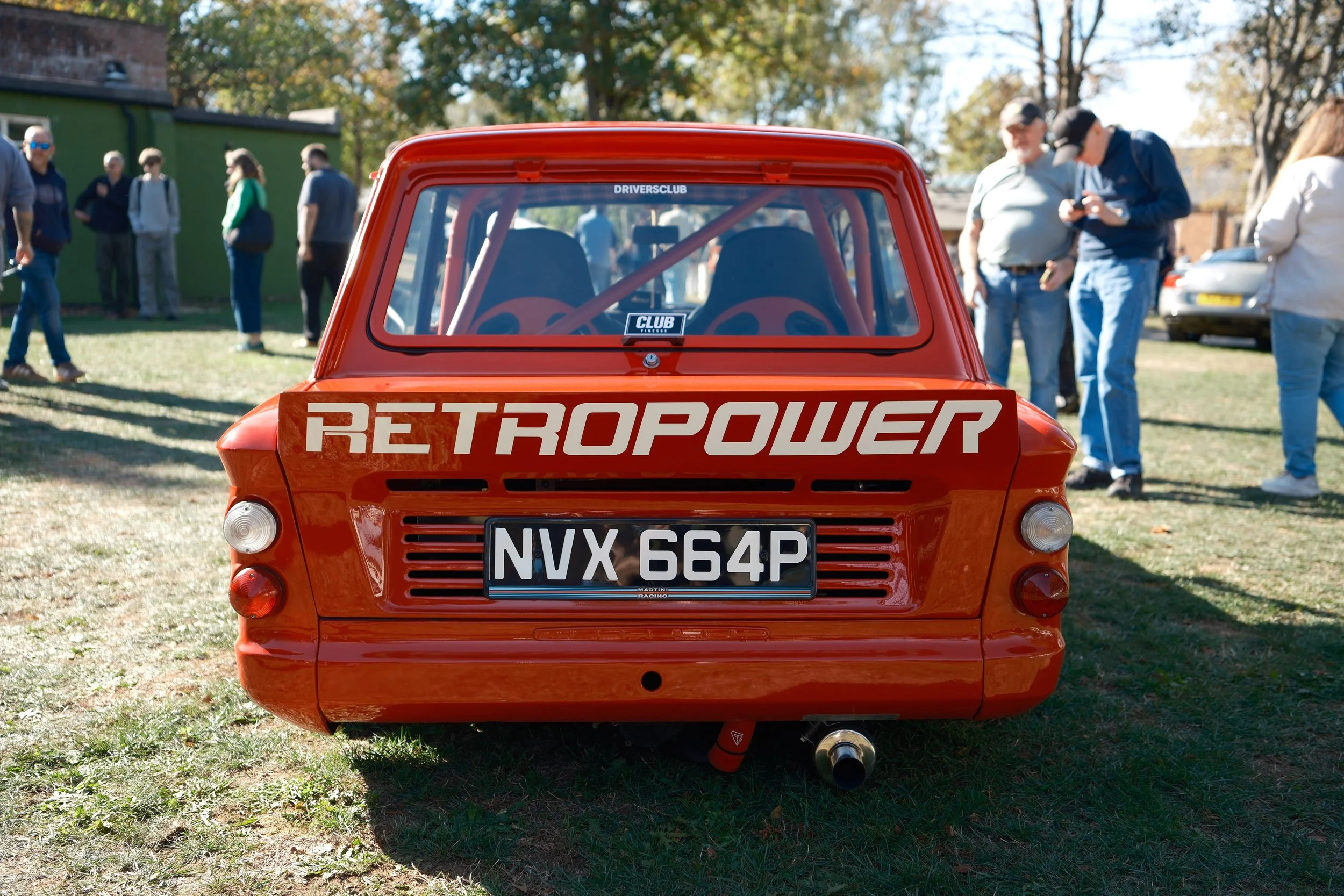 Back view of a red vintage Mini Cooper with rally-style racing modifications, including a roll cage inside, parked on grass during a car show, with people standing and conversing in the background.