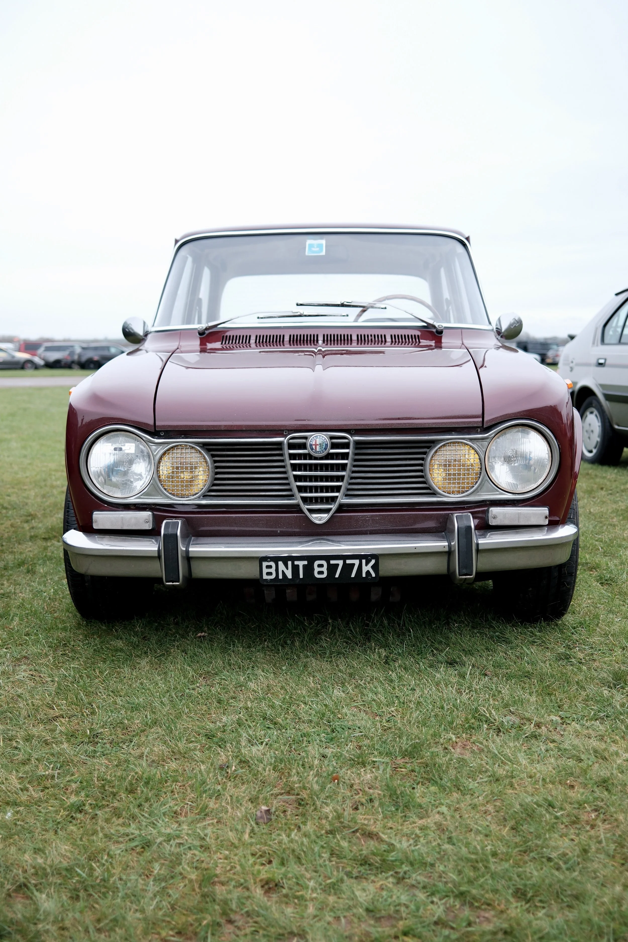 Front view of a vintage red car parked on grass, with other vehicles and a cloudy sky in the background.