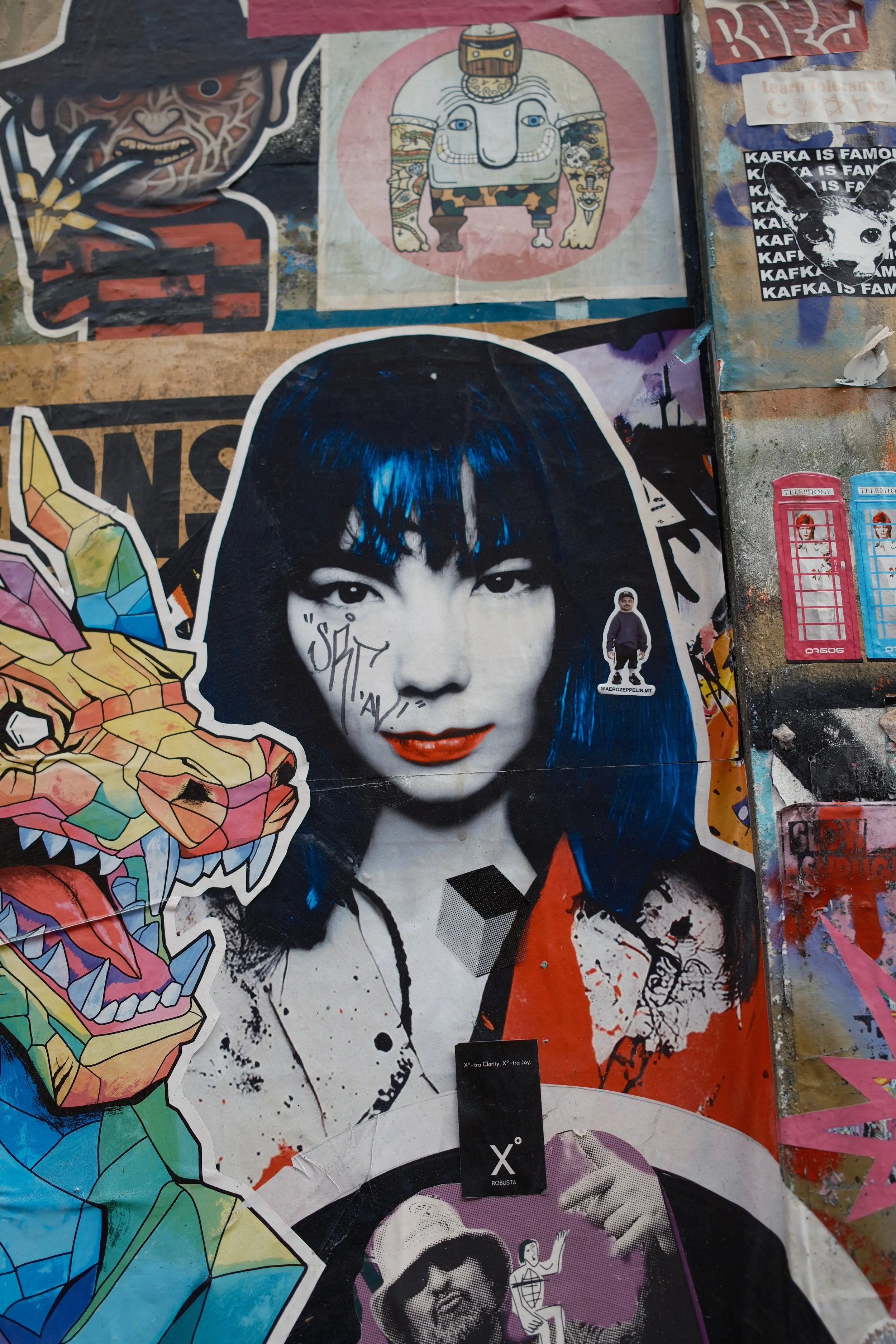 A collage of street art and stickers on a wall, featuring a black and white portrait of a woman with blue hair, various cartoon and graphic images, including a colorful dragon, a stylized computer, a dog with the text "KAFKA IS FAMOUS," and a small s