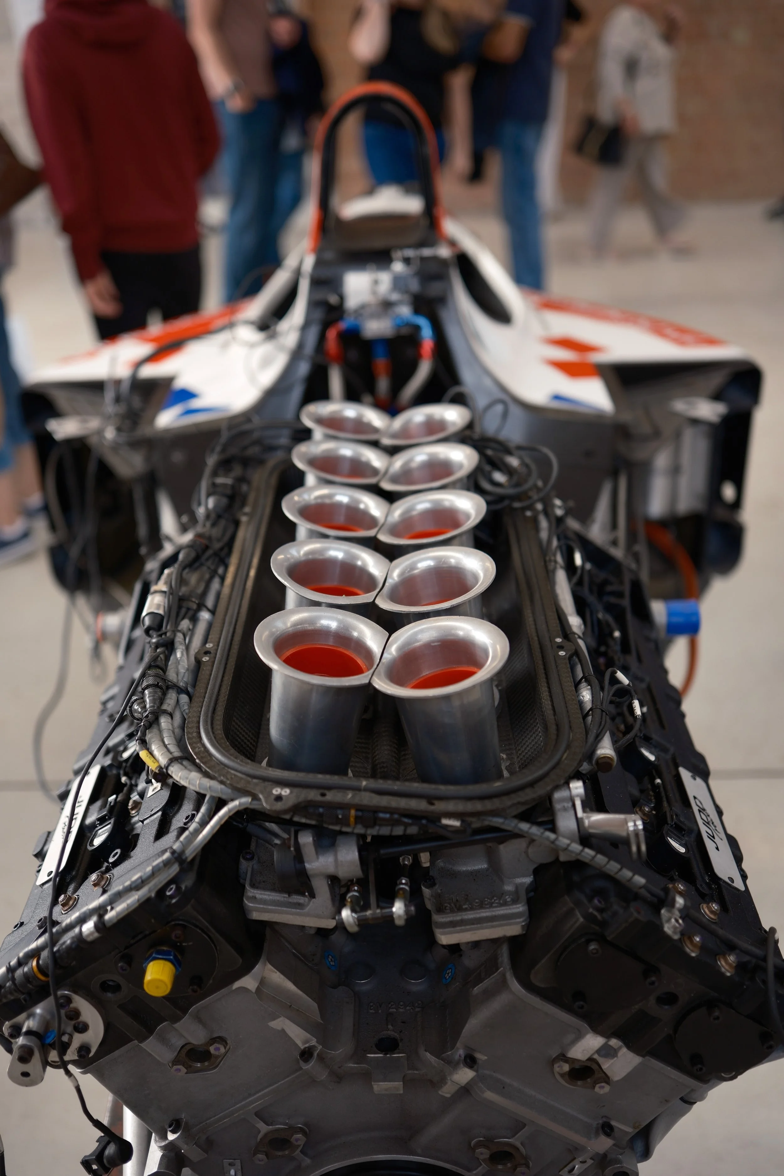 A racing car engine with multiple exhaust pipes, surrounded by wires and mechanical components, with a blurred group of people in the background.