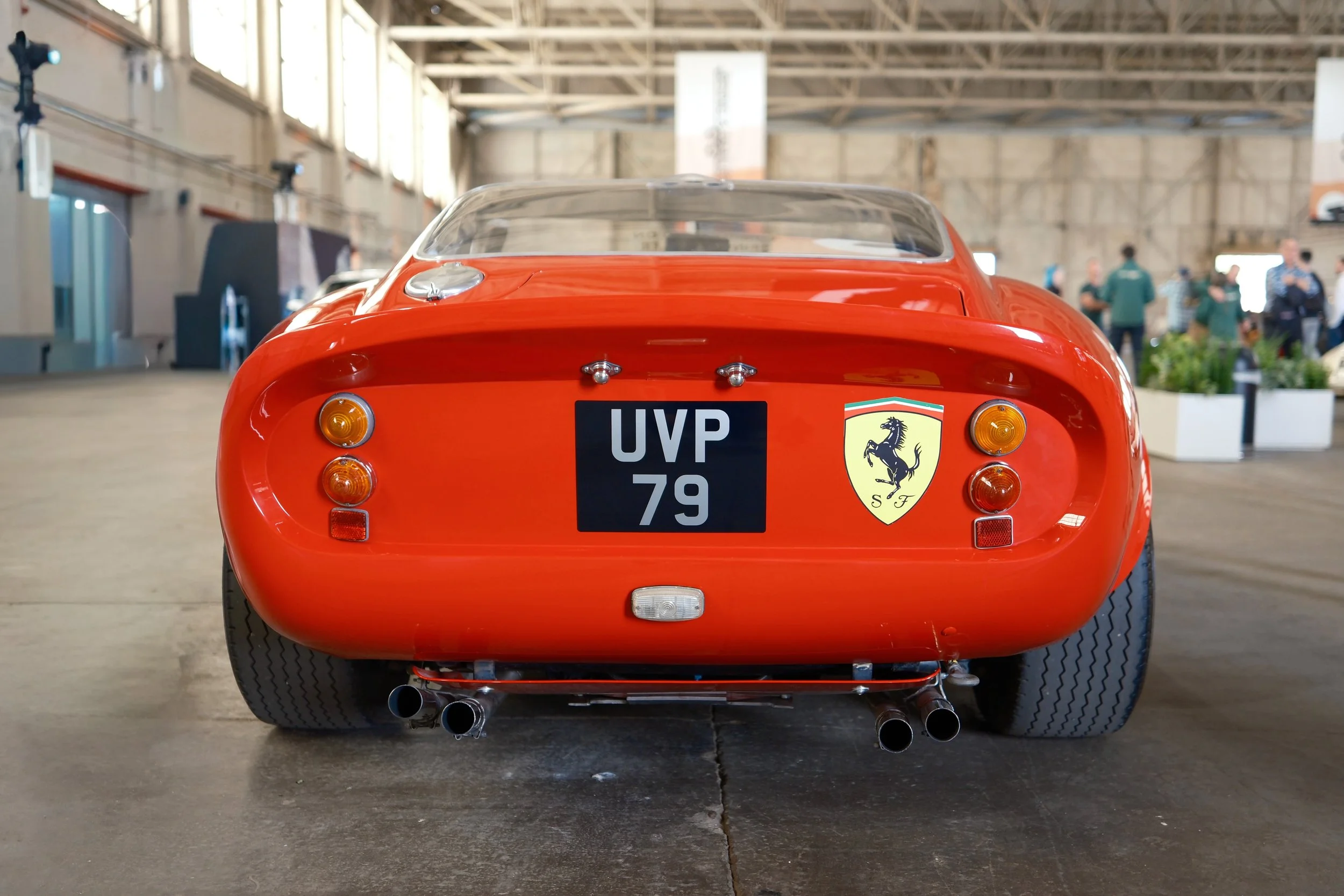 Rear view of a vintage red Ferrari race car displayed indoors, featuring the Ferrari logo, a black license plate with 'UVP 79', and four exhaust pipes.