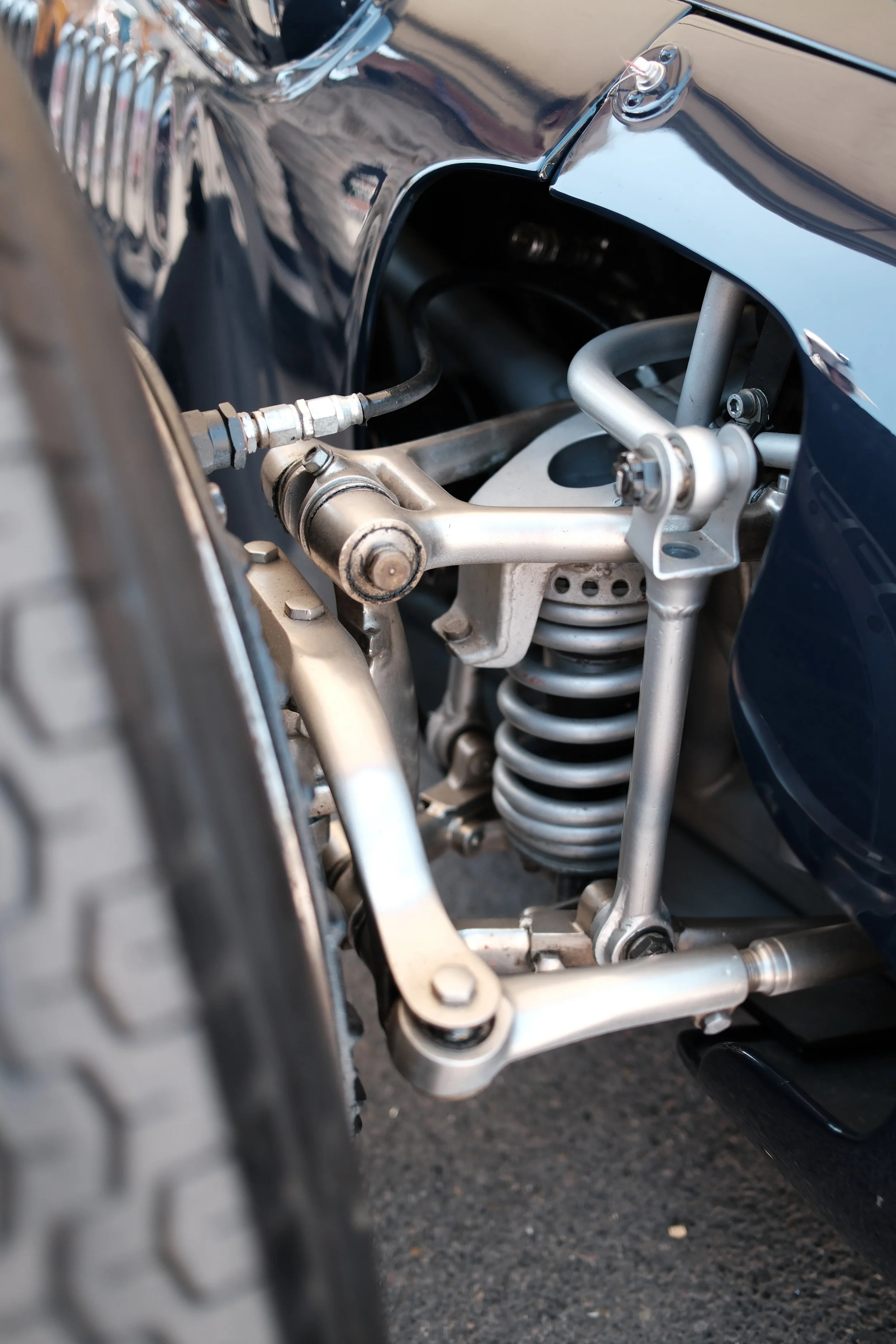 Close-up view of the suspension and wheel assembly of a vintage car, showing a coil spring, metallic linkages, and parts of the wheel tire in focus.