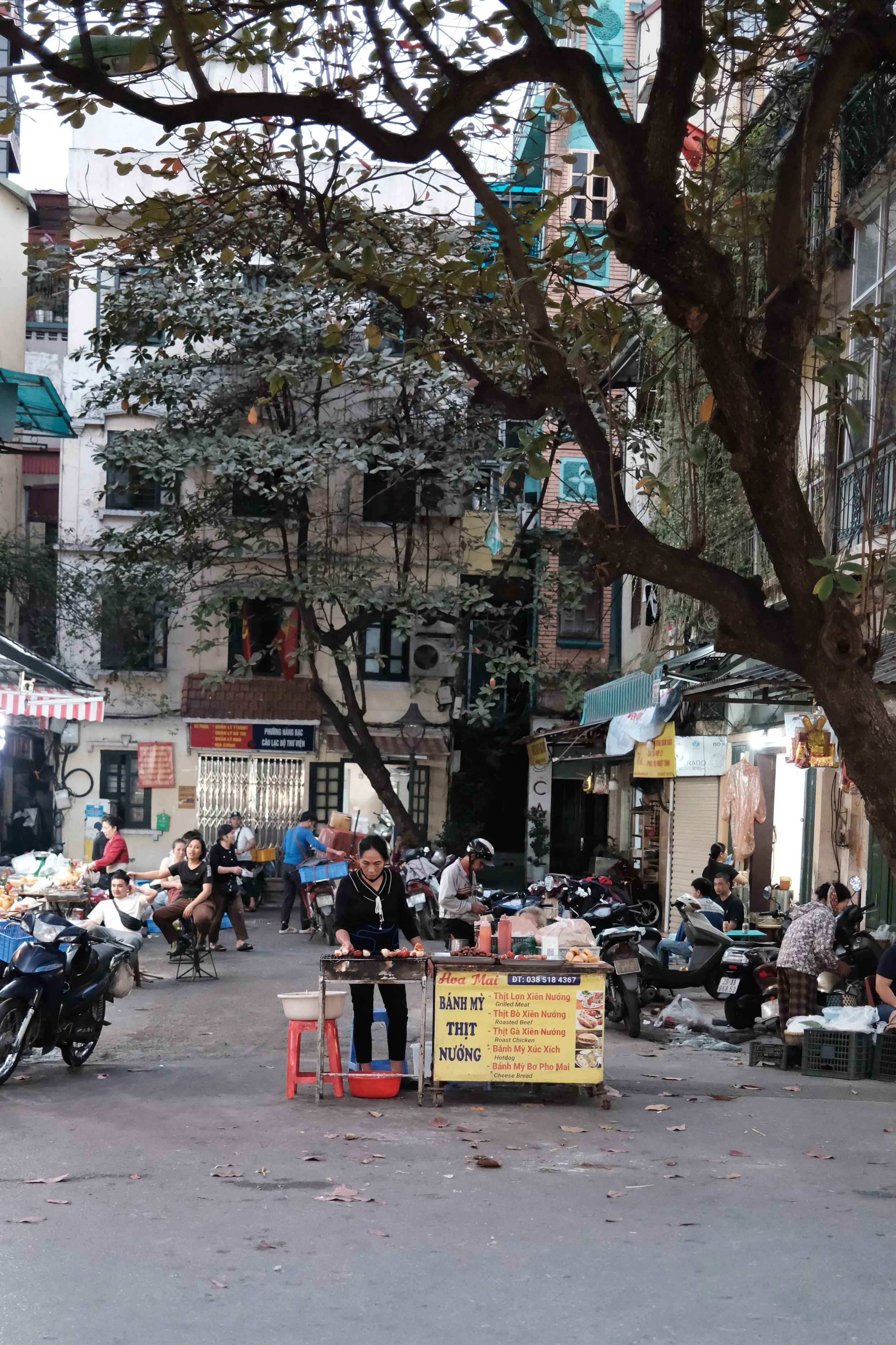 A busy street scene with multiple people, motorbikes, and street food vendors. There are signs with Vietnamese text and buildings in the background. The street is shaded by trees with leaves scattered on the ground.