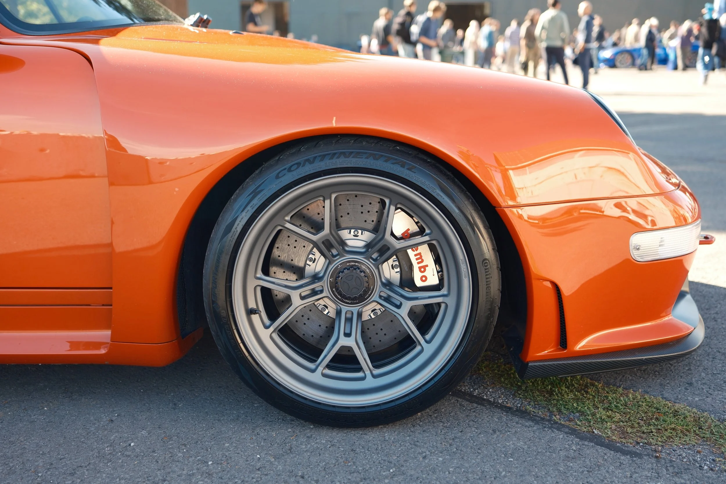 Close-up of an orange sports car front wheel with a Brembo brake system, parked on a street during a car show, with people in the background.