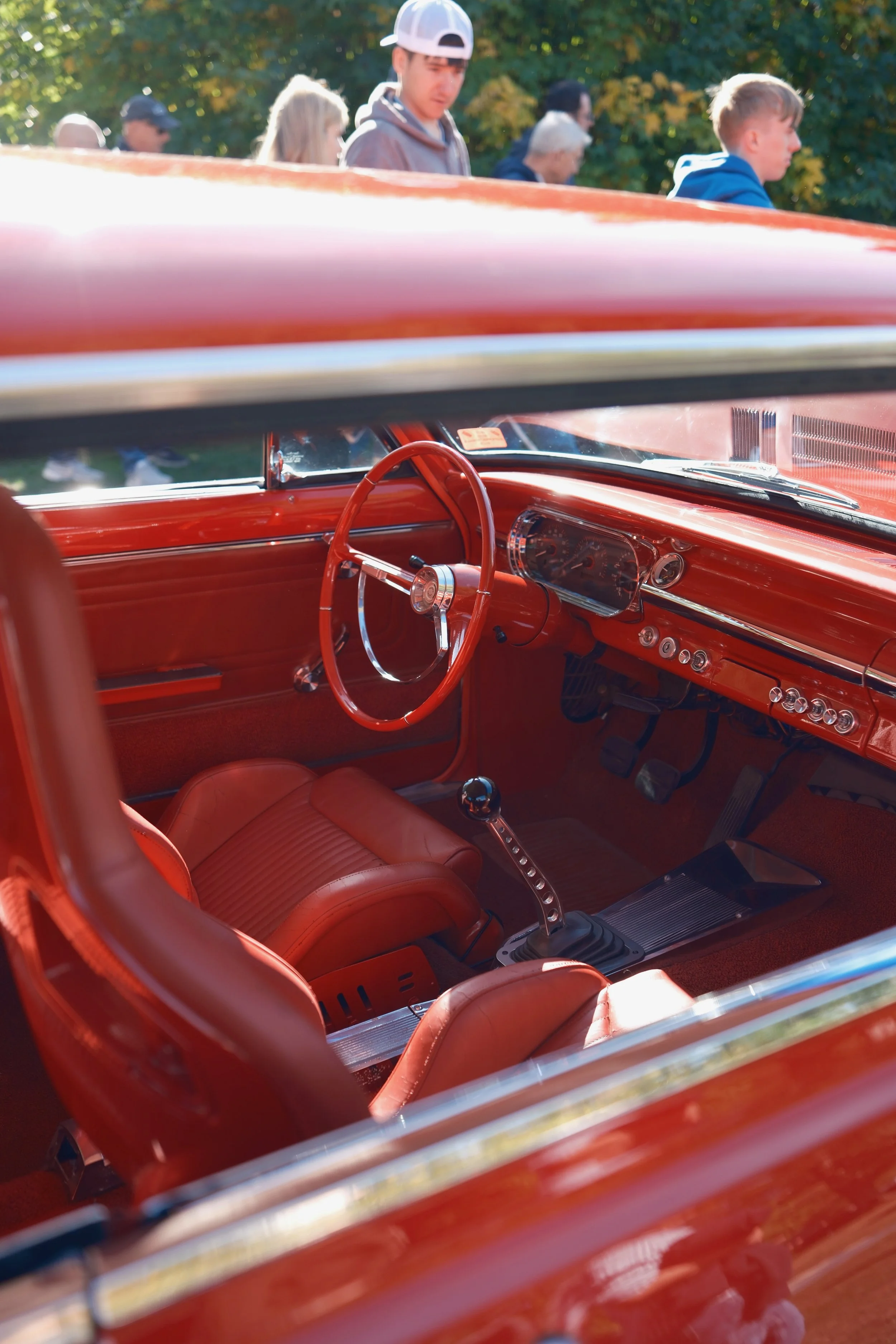 Inside view of a vintage red car with a red steering wheel, dashboard, and seats, taken from the passenger side, with a view of people walking outside through the windshield and side window.