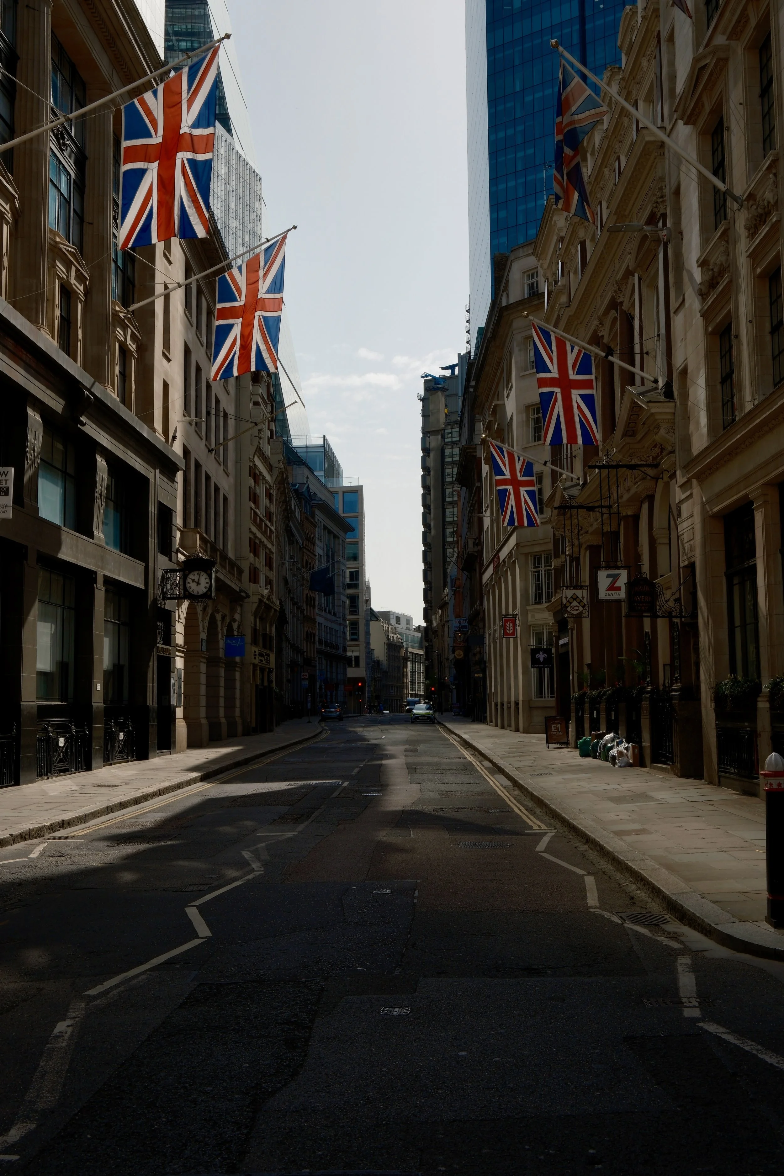 Empty city street with buildings on both sides and Union Jack flags hanging from the buildings. The street appears to be in London, with shadows cast on the pavement from nearby buildings. There are no pedestrians, and a few cars are parked or drivin