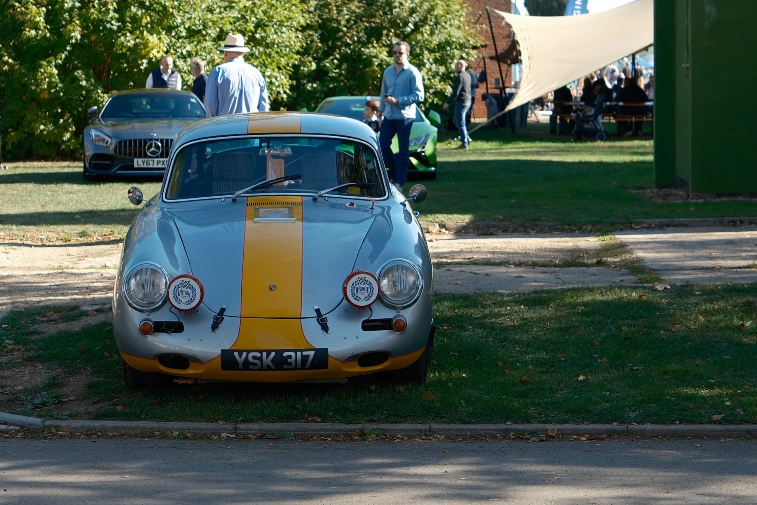A vintage silver race car with yellow racing stripe parked in a grassy area at an outdoor event, with luxury cars and people in the background.