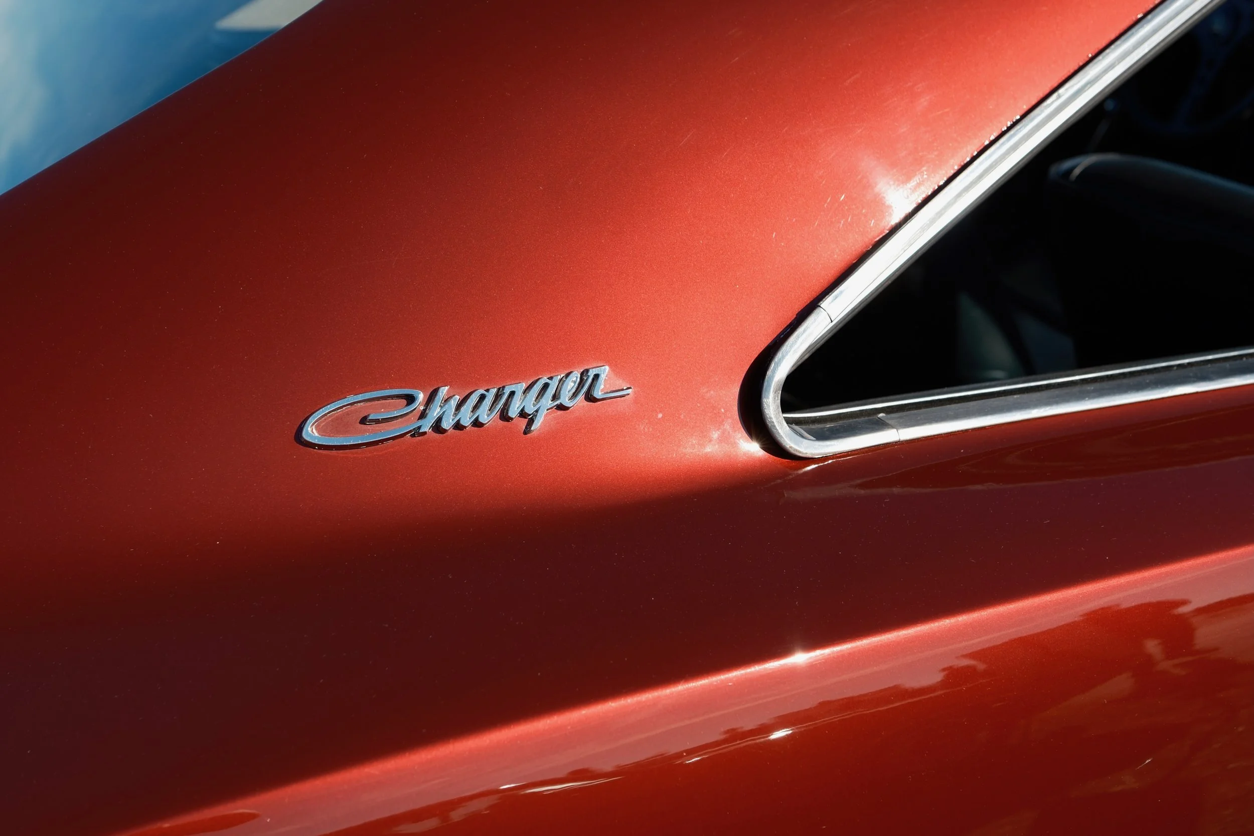 Close-up of a vintage car's red exterior with a chrome badge that reads 'Charger' and a vent. The car has a shiny, metallic finish.