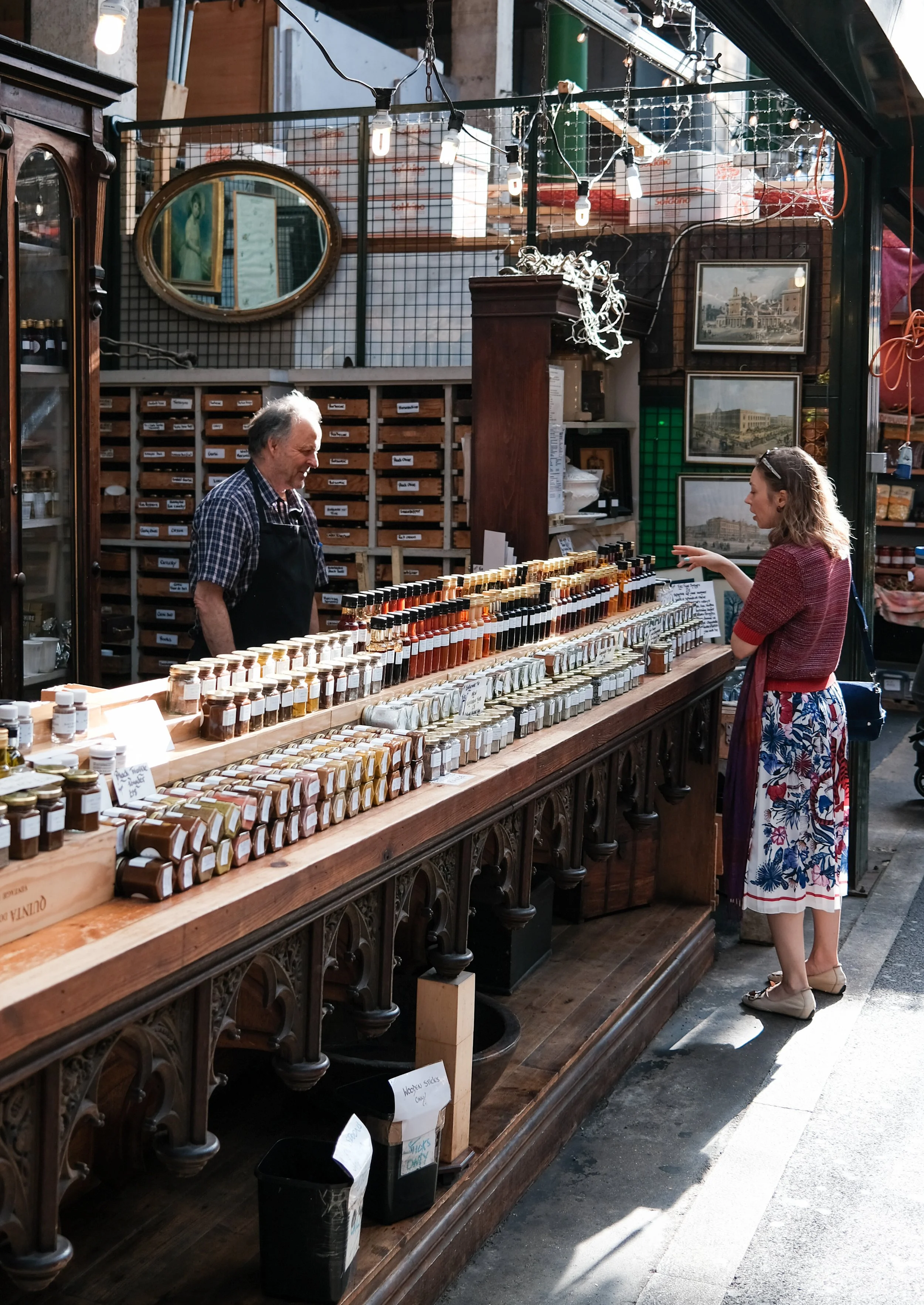 A woman browsing a spice stand at an outdoor market, talking to a vendor. The stand displays various spices in small jars, with framed pictures and a mirror hanging behind. The market appears to be vibrant, with sunlight illuminating the scene.